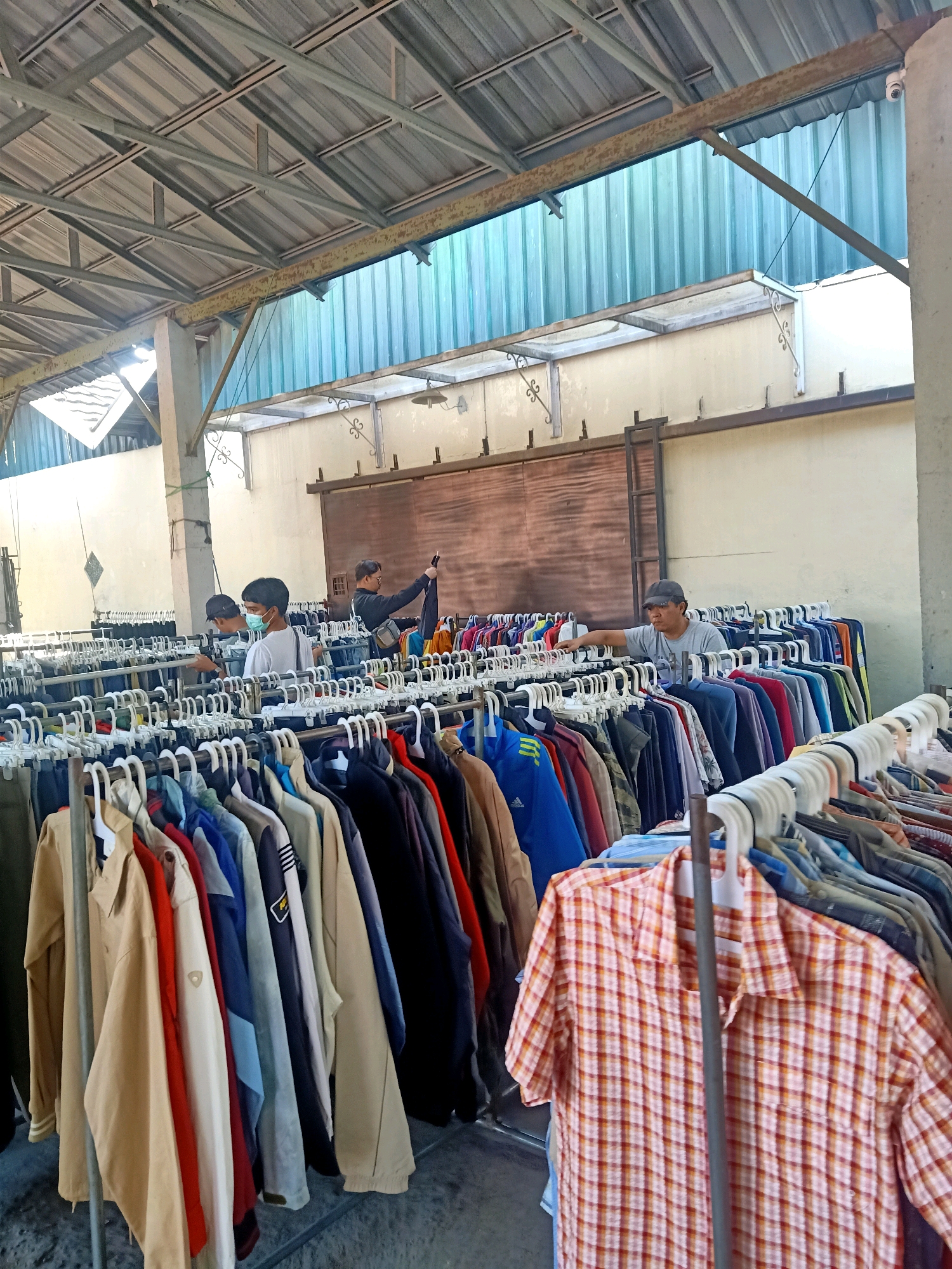 A street market stall is depicted in Jatinom, Indonesia, filled with racks of clothing for sale. The stall is covered by a corrugated metal roof supported by metal beams. Several people are browsing the clothes. In the foreground, a rack displays a variety of colorful shirts and jackets. Behind them, more racks of clothing are visible, with several individuals examining the merchandise. One person, wearing a surgical mask, is looking at shirts, while another man in a grey t-shirt and baseball cap is touching a rack of clothes. Further back, a man in a dark shirt is holding up a black garment. The background shows a wall and more metal roofing. The ground appears to be a concrete or dirt surface.