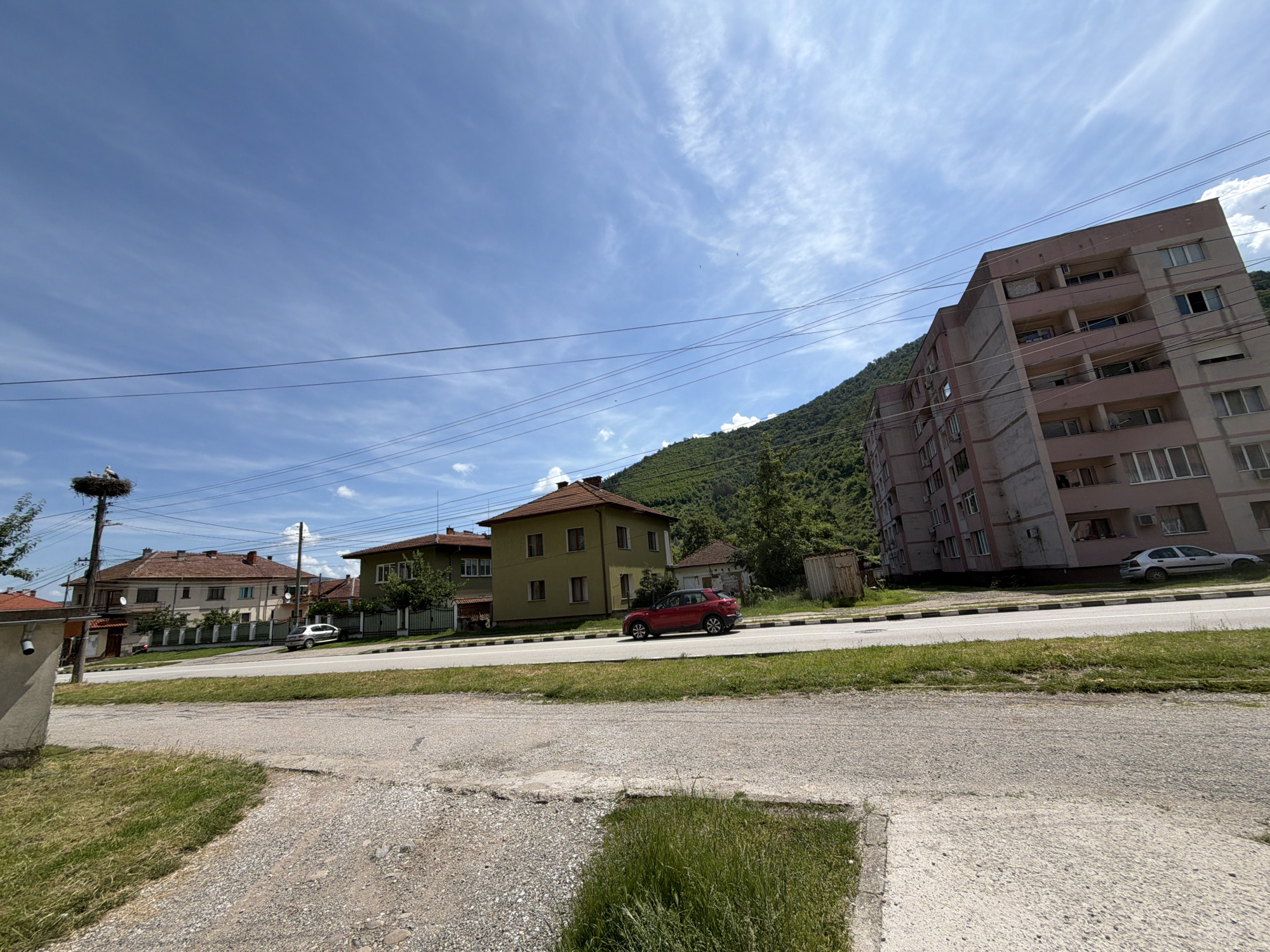 The image shows a street scene in Etropole, Bulgaria, on a sunny day with a blue sky and scattered clouds. In the foreground, there is a gravel and concrete area with grass growing through it. A paved road runs through the middle ground, with a white line marking the center and a black and white striped curb.
On the left side of the road, there are several houses. One is a light brown house with a red tile roof, and another is a green house with a red tile roof. A tall utility pole with a stork's nest on top is located to the left of the green house. Two storks are visible in the nest.
On the right side of the road, a large, pink and beige apartment building dominates the background. It has multiple floors with many windows. A small silver car is parked in front of this building. Further to the right, a lush green mountain slopes upwards, covered in trees.
A red SUV is driving on the road, moving away from the viewer. A white sedan is parked on the right side of the road, in front of the apartment building. The scene appears to be a quiet residential area.