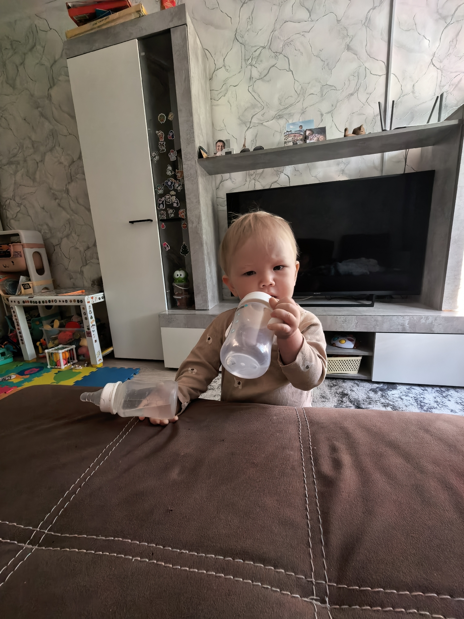 A wide-angle shot features a toddler drinking from a baby bottle in a room with a modern interior. The child has fair hair and skin, wearing a long-sleeved tan-colored outfit. They are holding one baby bottle to their mouth while another bottle lies on a brown couch. In the background, there is a white and gray entertainment center with a large flat-screen television. To the left, a white cabinet is adorned with stickers and a small white table with colorful toys. The walls are decorated with a textured, marbled pattern. The location is Sterlitamak, Russia.