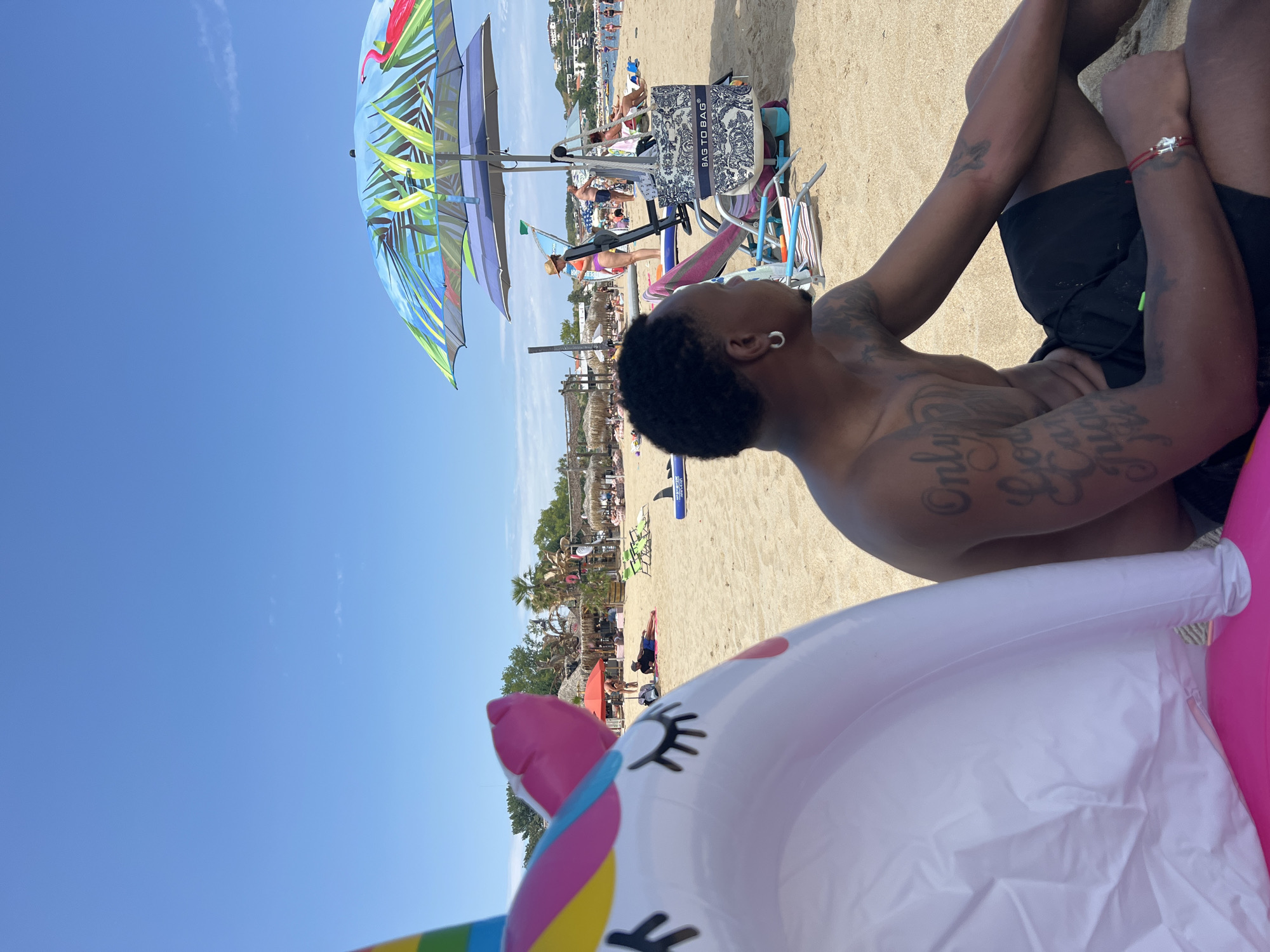 A man with tattoos on his arms and chest sits on a sandy beach in Sozopol, Bulgaria. He is wearing black swim trunks and an earring. A large, inflatable unicorn float is partially in the foreground. In the background, there are beach umbrellas, beach chairs, and people relaxing on the sand. The sky is blue with some light clouds.