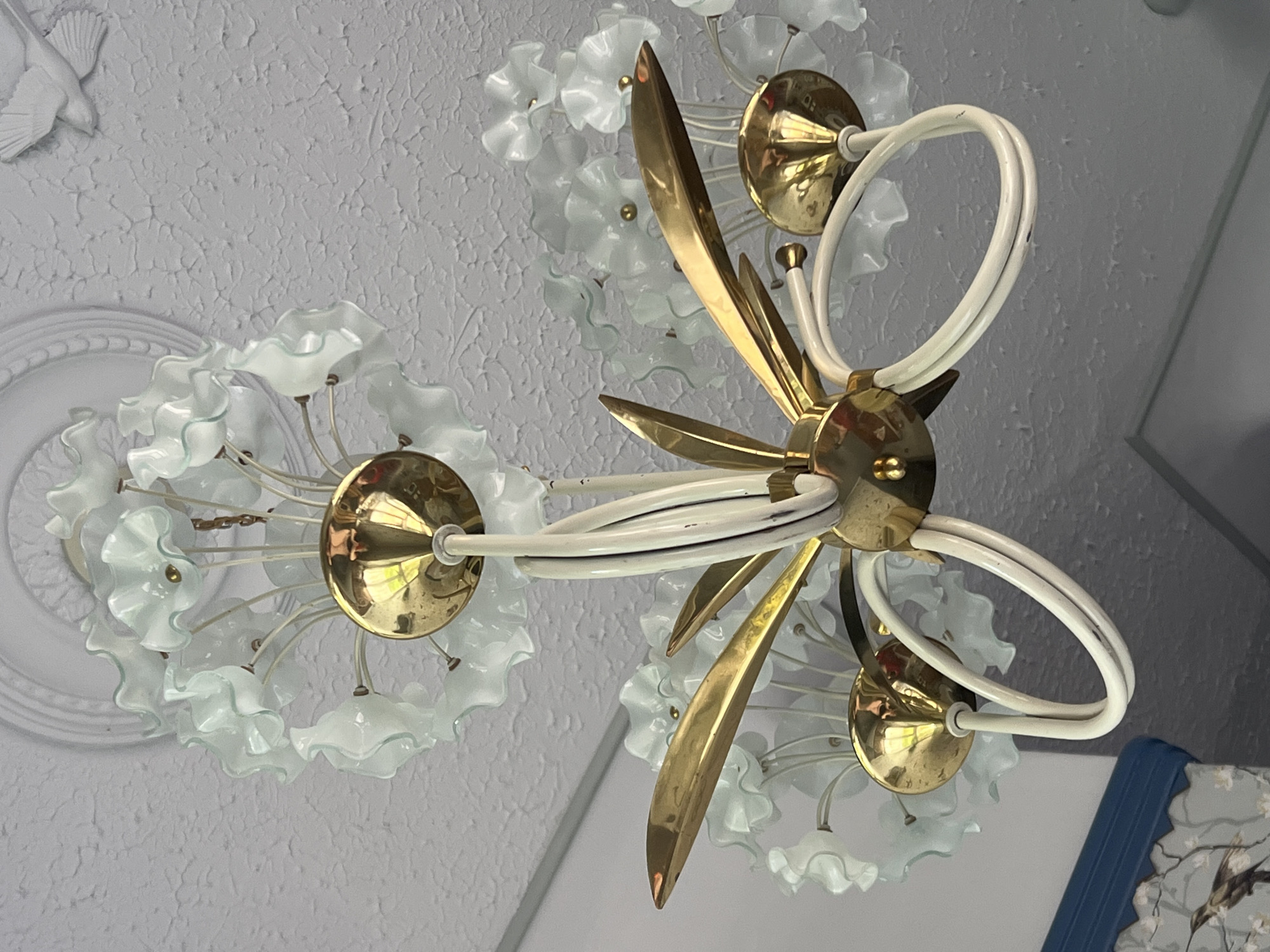 The image shows a close-up view of a vintage chandelier hanging from a textured white ceiling. The chandelier features a central brass fixture with white, curved arms that extend outwards. It is adorned with numerous delicate, translucent glass elements that resemble ruffled petals in a pale blue or frosted white hue. Some of these glass elements have small brass caps. The ceiling itself has a decorative relief molding around it, and a white bird figurine is mounted on the wall in the upper right corner of the frame. A portion of a wall with a blue trim and wallpaper featuring a bird and floral motif is visible in the lower left. The scene appears to be an indoor setting, possibly a home or a decorative space. There is no indication of the city of Veliky Novgorod, Russia, in this image.