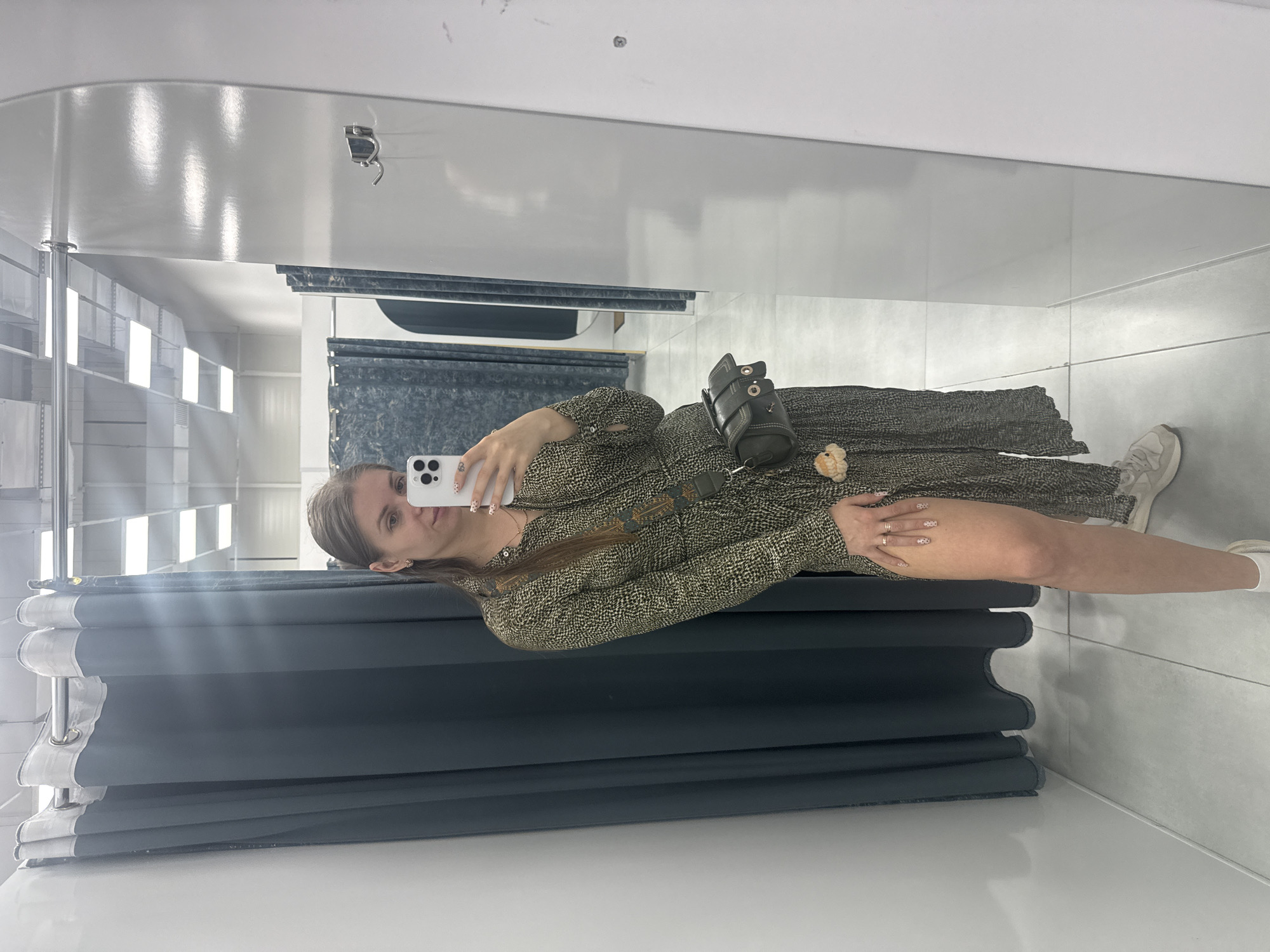 The image depicts a young woman taking a selfie in a dressing room. She is wearing a patterned dress, a crossbody bag, and sneakers. The dressing room has gray curtains and white walls. The location is likely a retail store in Batumi, Georgia, based on the architectural style and general aesthetic.