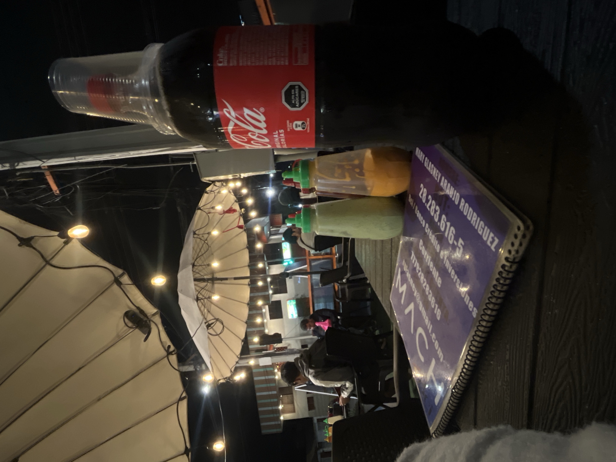 This is a nighttime, medium-shot, eye-level photograph taken at a casual outdoor dining establishment in Iquique, Chile. The scene is dominated by a large bottle of Coca-Cola positioned prominently in the foreground to the right. The bottle has a clear plastic cup covering its opening. To the left of the Coca-Cola bottle are two condiment squeeze bottles, one with a pale green liquid and the other with an orange-yellow liquid. A menu, with purple and white text, lies open on a dark wooden table in the foreground, partially obscured by a person's gray sleeve on the left side of the frame.
In the middle ground, several people are seated at tables. One person in a gray and black jacket is looking down at something on their table, while another person in the background wears a pink beanie. The establishment appears to be set up with several large umbrellas providing shelter, illuminated by strings of warm-toned fairy lights overhead. In the background, there are buildings with storefronts, some with visible screens or lights. The overall ambiance suggests a relaxed, open-air eatery.