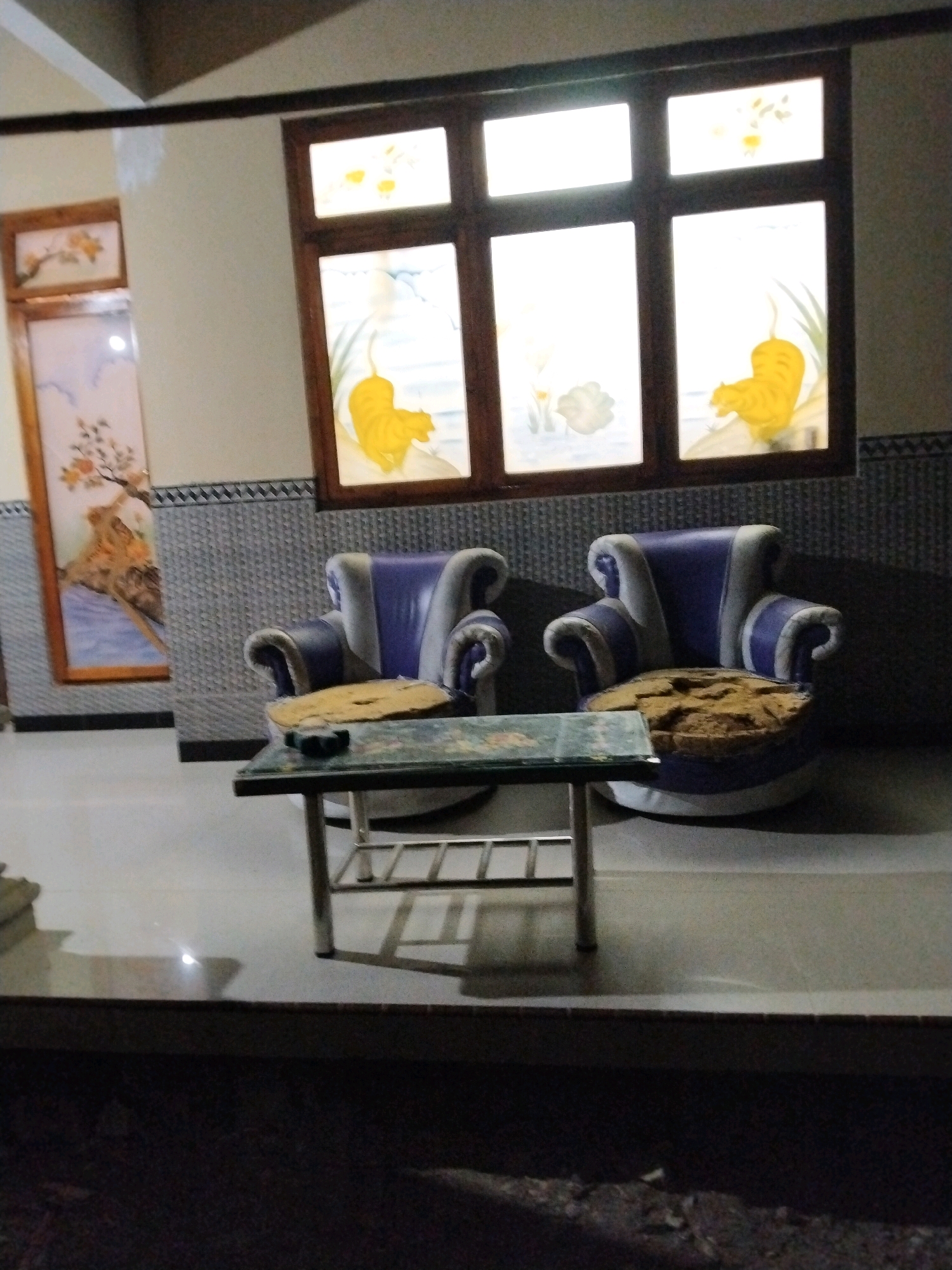 The image shows a dimly lit indoor scene, likely a living room, with two plush armchairs and a coffee table. The armchairs are upholstered in a combination of purple and white, with large, rolled arms. Both chairs appear to be worn, with the cushions looking flattened and aged. The coffee table is rectangular with a glass top and a metal frame. A small black object, possibly an ashtray, sits on the table. Behind the furniture, a large window with multiple panes features decorative stained glass. The stained glass depicts yellow tiger-like figures and floral motifs. To the left of the window, a framed artwork hangs on the wall, displaying a scenic landscape with a tree. The walls are covered with patterned tiles in shades of blue and grey. The flooring appears to be tiled. The overall impression is one of a domestic setting, possibly an older establishment or a space decorated with traditional elements. The lighting is dim, casting shadows and suggesting it might be evening or the room is not brightly illuminated. Without additional context, a specific location within Tasikmadu, Indonesia, cannot be determined from the image alone.