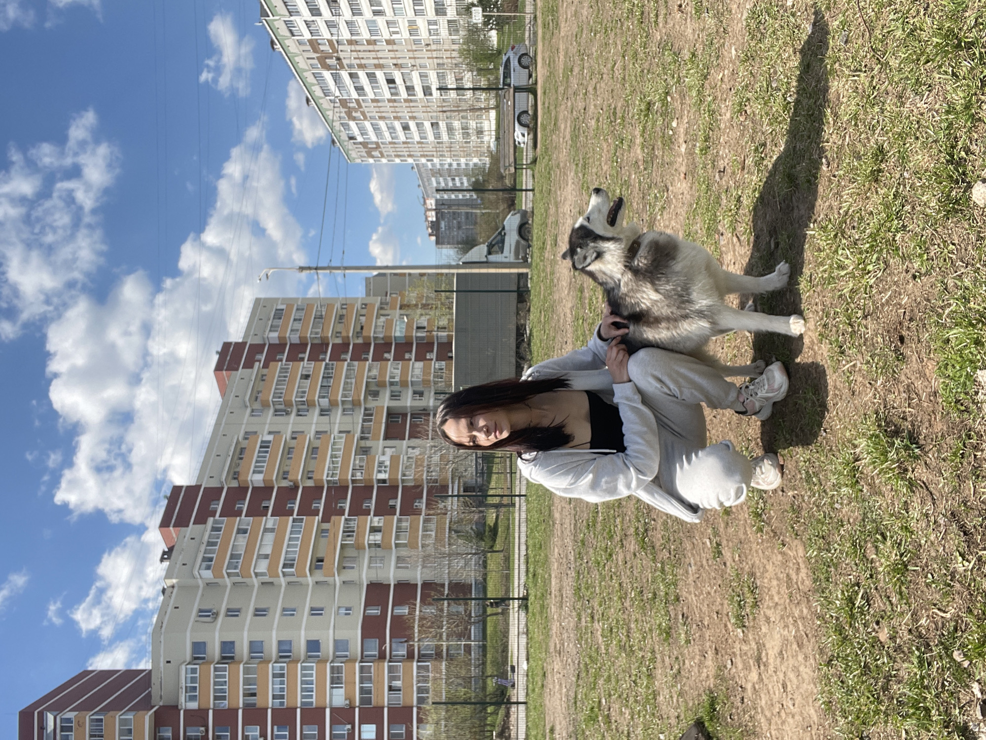A woman and a husky dog are in a park in Izhevsk, Russia, on a sunny day. The woman is crouching, wearing a grey tracksuit and white sneakers. She is holding the dog, which is standing and looking up. In the background are apartment buildings and a blue sky with clouds. A fence is visible behind the woman. The ground is a mix of grass and dirt.
