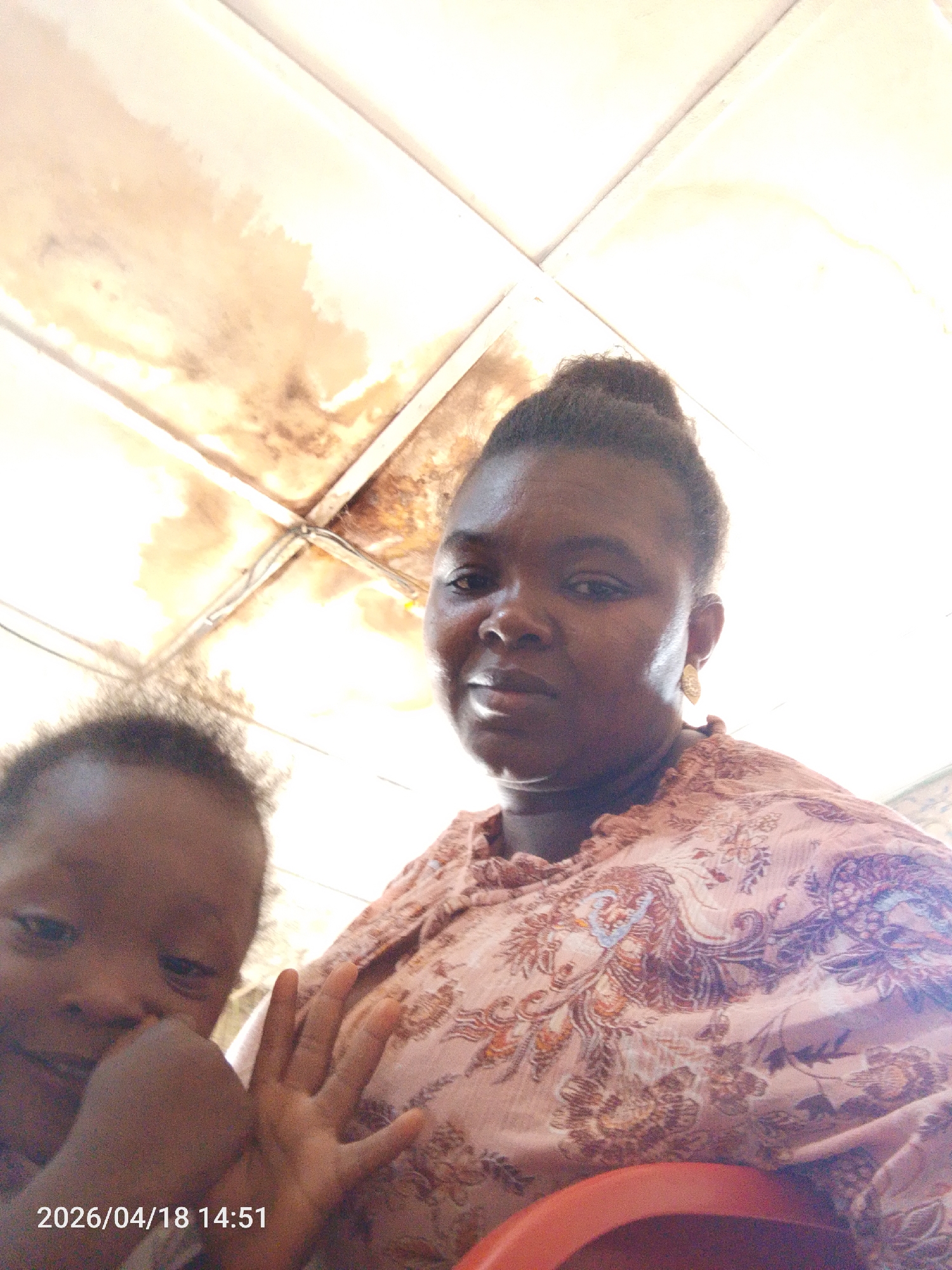 A low-angle shot shows a woman with a child in what appears to be an indoor setting. The woman is dark-skinned with her hair in a bun, wearing a patterned light pink top and dangling earrings. To her left, a child is partially visible, holding up a hand. The background shows a weathered, possibly water-damaged ceiling with a grid pattern. The location, Lokoja, Nigeria, is inferred as the origin. A date stamp "2026/04/18 14:51" is present at the bottom left corner of the image.