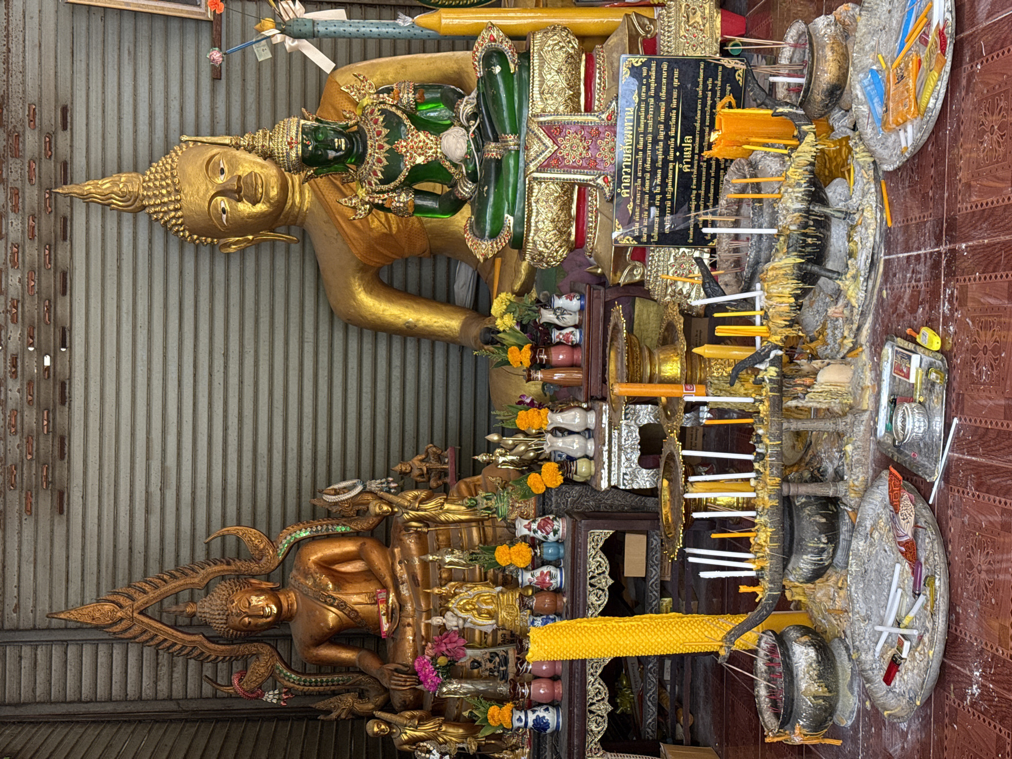 The image depicts an outdoor shrine featuring two large Buddha statues and numerous offerings. The primary subject is a large, gold-colored Buddha statue seated on a raised platform, with a smaller, green Buddha statue placed in front of it. Both statues are adorned with intricate golden crowns and elaborate jewelry.
In front of the statues, a tiered wooden shelf displays an array of small vases, some containing flowers, and decorative items. Below this shelf, a collection of unlit candles of various sizes, incense sticks in burners, and trays holding what appear to be offerings such as small packets and potentially matches are arranged on the floor. The ground is tiled with a red patterned material.
The background consists of a weathered, corrugated metal roll-up door, suggesting the shrine is located in a commercial or storage area. The overall scene is illuminated by natural light. The presence of multiple unlit candles, incense, and offerings indicates this is a place of worship or remembrance. The context of Ko Samui District, Thailand, suggests this is a Buddhist shrine, common in such locations.