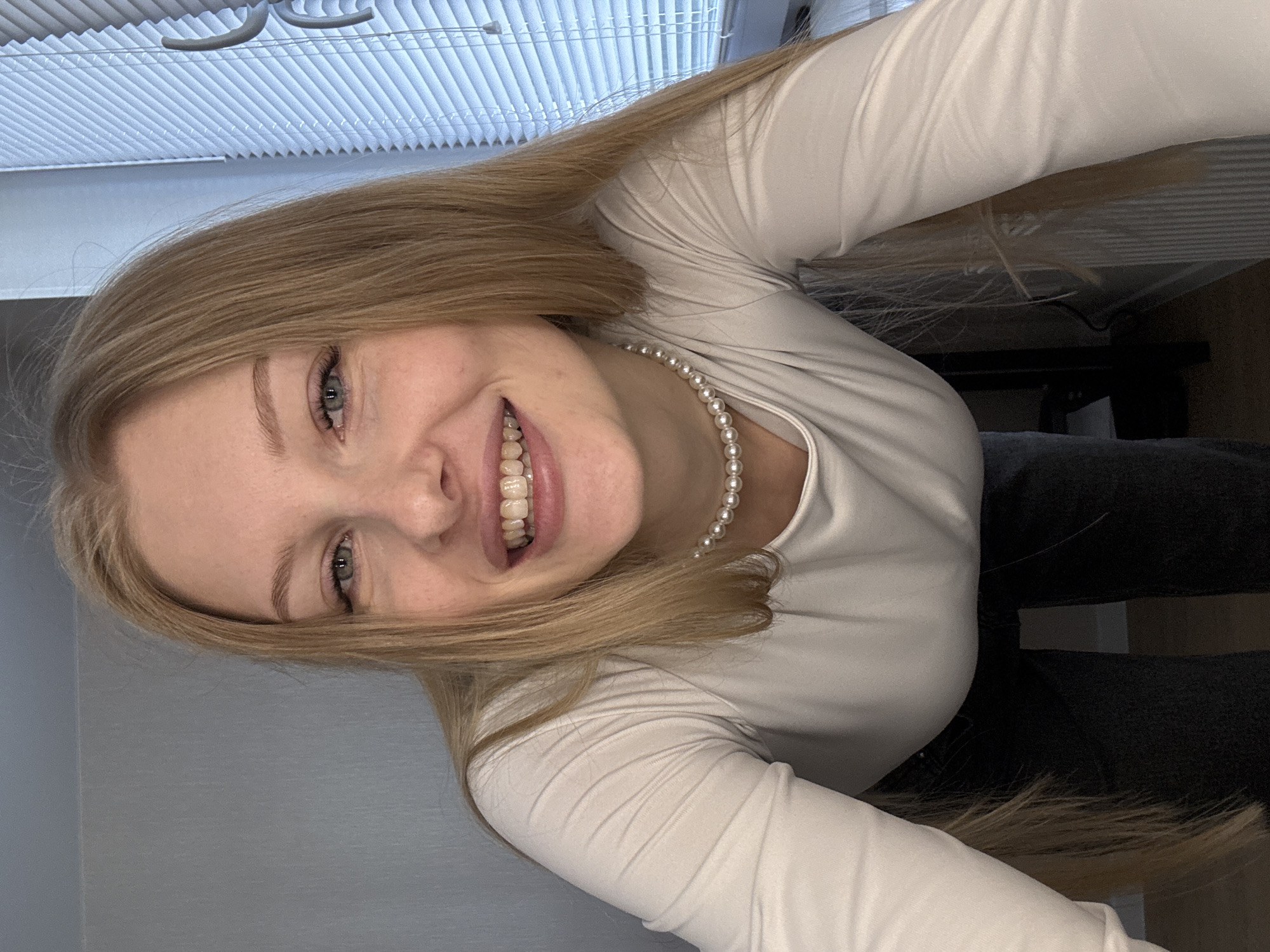 This media file features an angled indoor portrait of a young woman, likely a selfie, taken in Perm, Russia. The woman has long, straight blonde hair and fair skin. She is smiling broadly, revealing her upper teeth, and her eyes are a light blue or grey color, complementing her neatly shaped light eyebrows. She wears a simple, light-colored, long-sleeved top, possibly white or off-white, paired with a classic pearl necklace. A portion of dark clothing, perhaps trousers, is visible in the lower right of the frame.
The background shows elements of an indoor setting. White horizontal window blinds are visible in the upper part of the image, suggesting a window is nearby. Above these blinds, some grey, curved fixtures can be seen, possibly part of the ceiling or a lighting unit. To the left, a plain light grey wall serves as a backdrop, while the lower right includes dark, indistinct shapes that appear to be furniture or fabric. The lighting is soft and consistent, highlighting her features. The woman's direct gaze and cheerful smile convey a happy and engaging emotion. No visible text is present in the image.