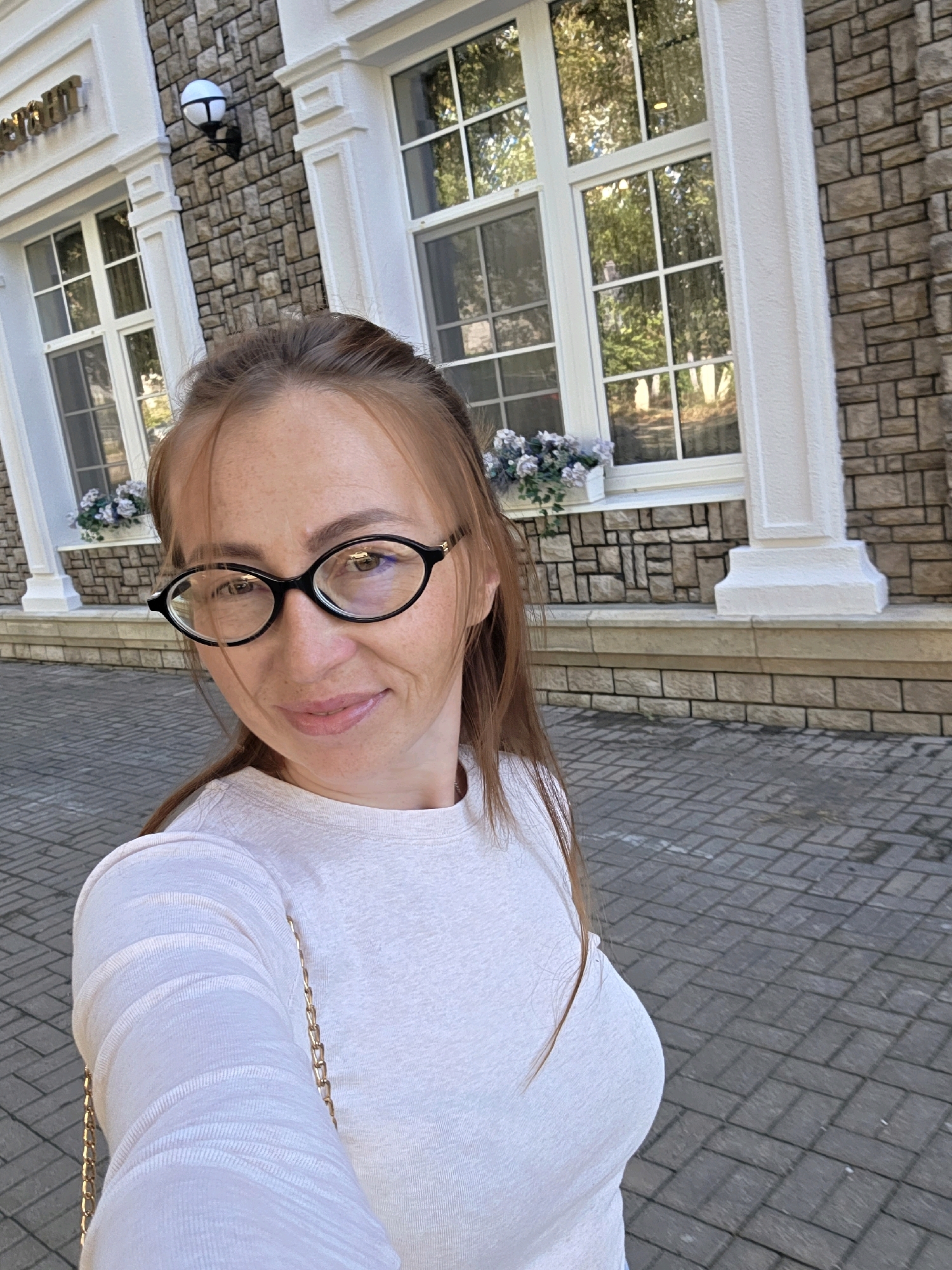 A medium shot photograph captures a woman with light brown hair, wearing glasses and a white long-sleeved shirt, posing for a selfie outdoors. She is positioned in front of a building with a stone facade and large windows. The windows reflect trees and sky, suggesting it is daytime. There are decorative planters with flowers below the windows. The ground is paved with gray interlocking bricks. The overall scene appears to be in a well-maintained urban environment. The text on the building, partially visible, appears to be "ресторан" (restaurant in Russian). The building's architecture and the presence of the word "ресторан" suggest a location in a city. Without further information, it is impossible to definitively place this image in Votkinsk, Russia, as similar architectural styles and establishments can be found in many Russian cities.