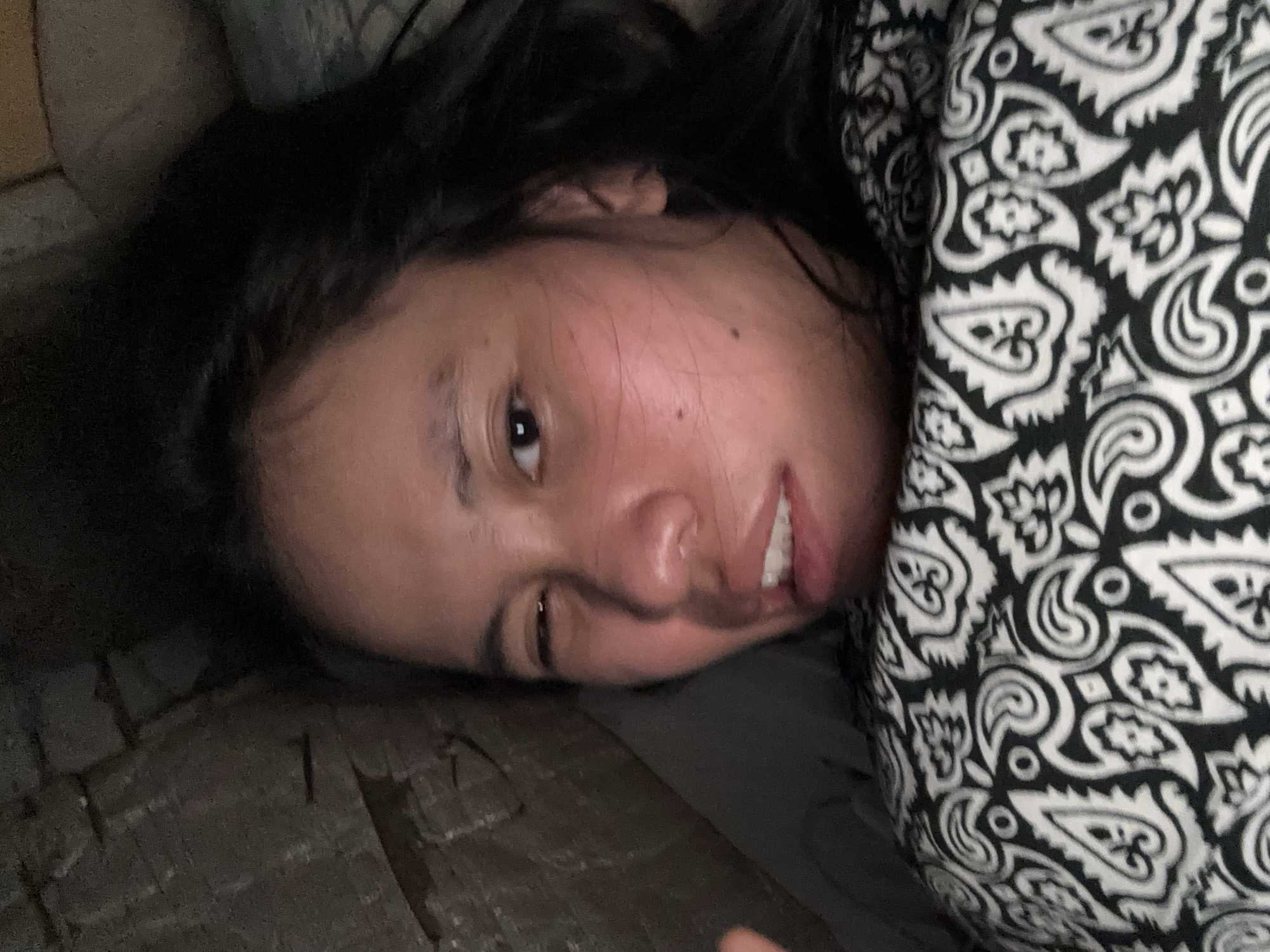 A person is lying down with their head propped up against a rough, brick-like surface. Their body is covered with a black and white patterned fabric. The person's face is close to the camera, with one eye open and the other partially closed. Their mouth is slightly open, revealing teeth. The lighting is dim, casting shadows on the face. The background appears to be an indoor environment with a textured wall.
