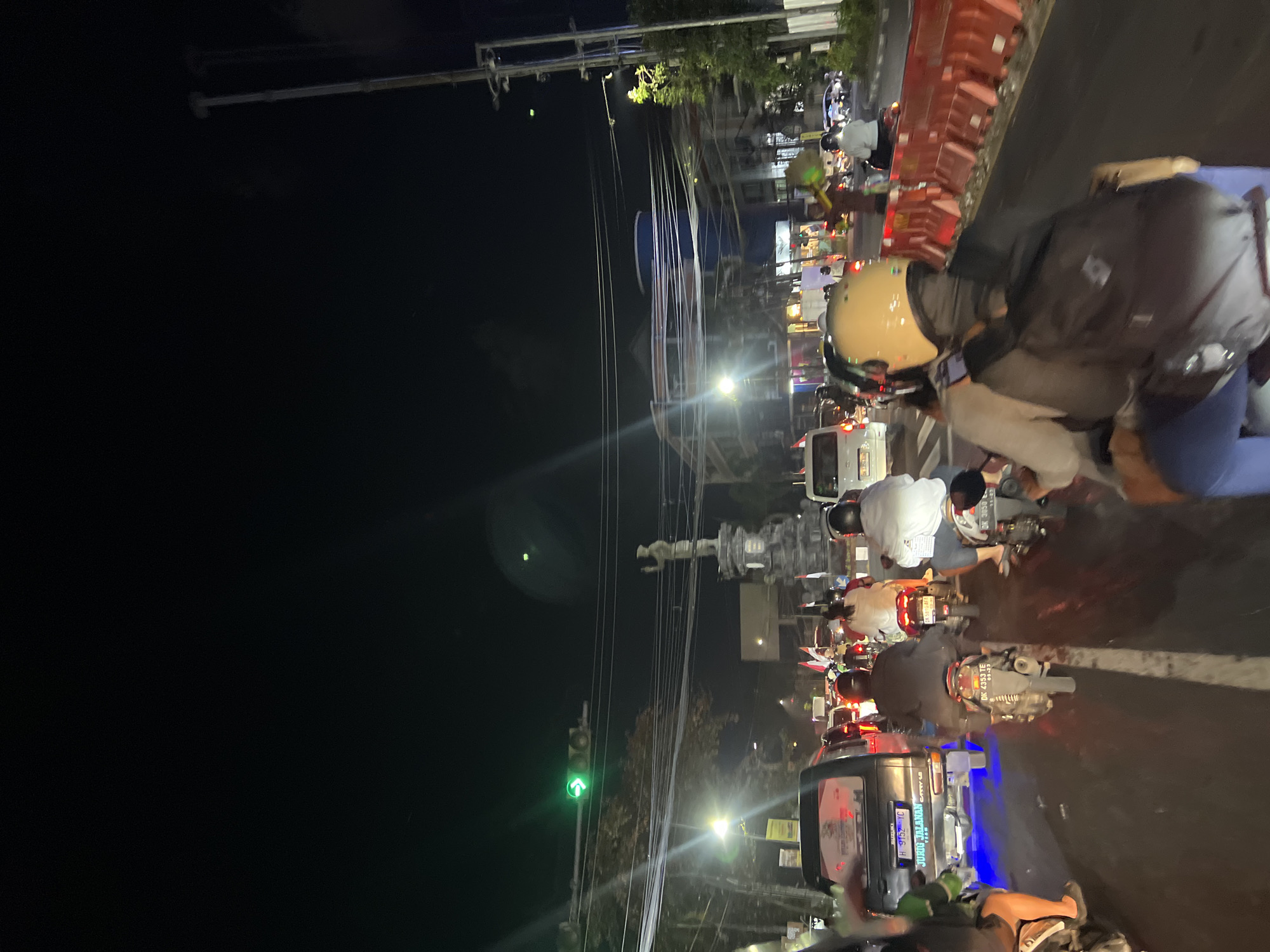 The image captures a nighttime street scene in Denpasar, Indonesia, characterized by heavy traffic. Several motorcycles and cars are visible, navigating a busy road. A traffic light shows a green arrow, indicating permission to proceed in that direction. In the background, a monument stands tall, illuminated by streetlights. The environment is urban, with buildings and utility poles lining the street. The overall atmosphere suggests a typical evening commute or event in the city, with people on motorcycles and in vehicles. The presence of flags further suggests a possible celebration or public gathering. The road surface appears wet, possibly from recent rain, reflecting the surrounding lights.