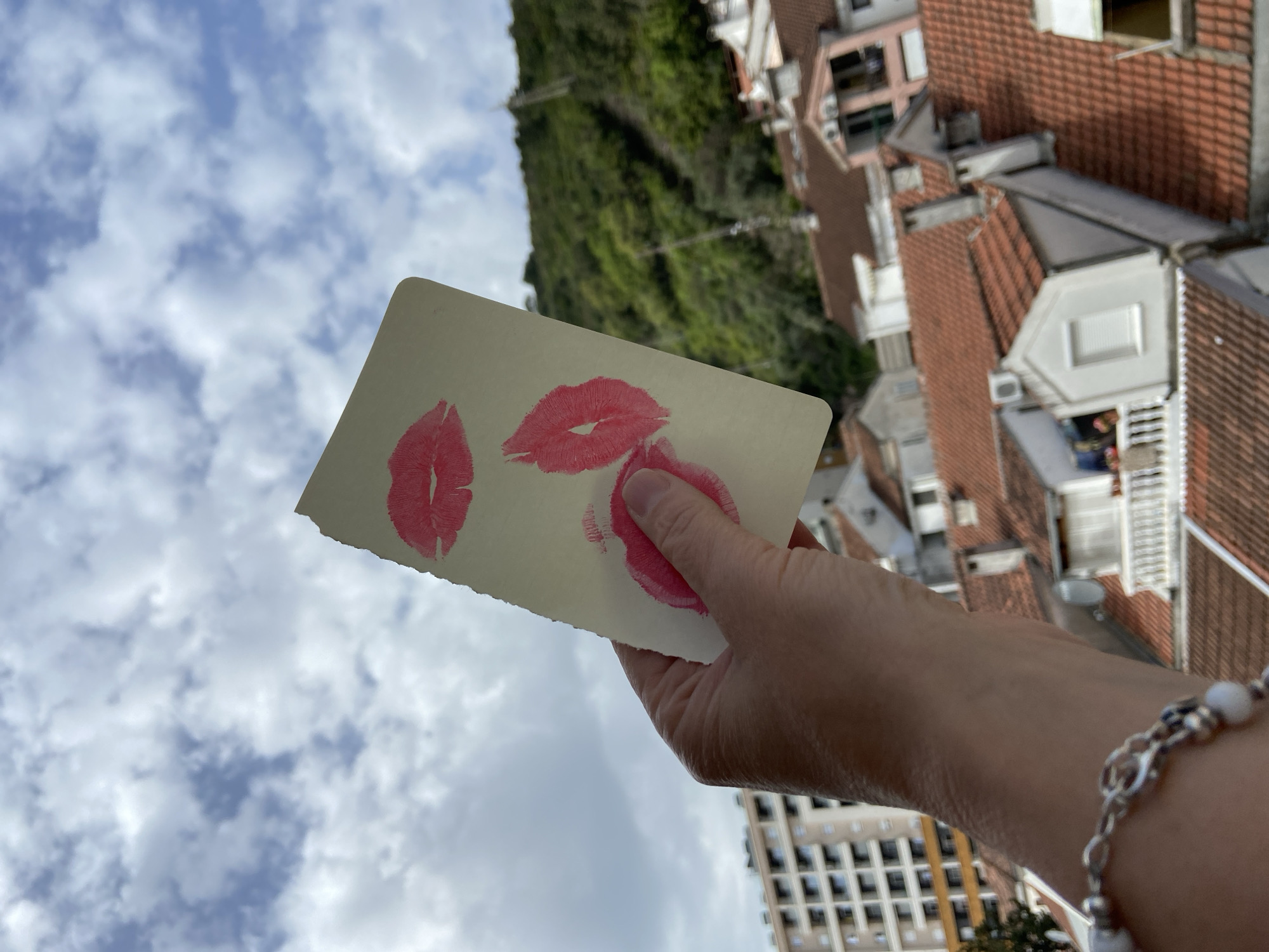 A medium-shot, tilted photograph depicts a hand holding a rectangular piece of paper with three lipstick kiss imprints against a backdrop of the cloudy sky, buildings, and greenery. The sky fills the lower-left quadrant of the scene, displaying white clouds against a blue expanse. The hand, adorned with a beaded bracelet, is positioned near the bottom-right corner, displaying the card with the lipstick marks. The card shows three distinct imprints of lipstick. Above the card, a cluster of buildings with red-tiled roofs is visible, typical of the architecture found in old European cities. Beyond the buildings, a hillside covered in dense green vegetation stretches across the upper portion of the image. A tall building can be seen between the arm and the sky. The scene is a cityscape, likely from an elevated position. The city location is Budva, Montenegro, as indicated by the building style and landscape characteristics.