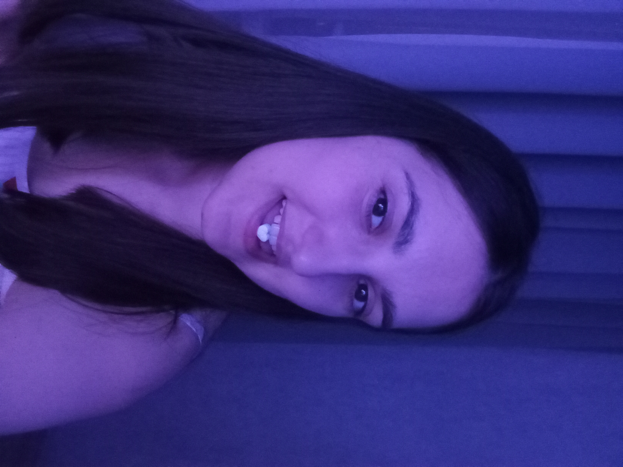 A young woman with dark, straight hair is captured in a close-up portrait. She is looking at the camera with a slight smile, revealing a small, white object, possibly a pill or candy, resting on her teeth. The lighting in the image is predominantly purple, casting a soft glow on her face and the blue curtains in the background. The woman appears to be indoors, in a dimly lit room. No specific geographical features identifiable for Krasnodar, Russia are present.