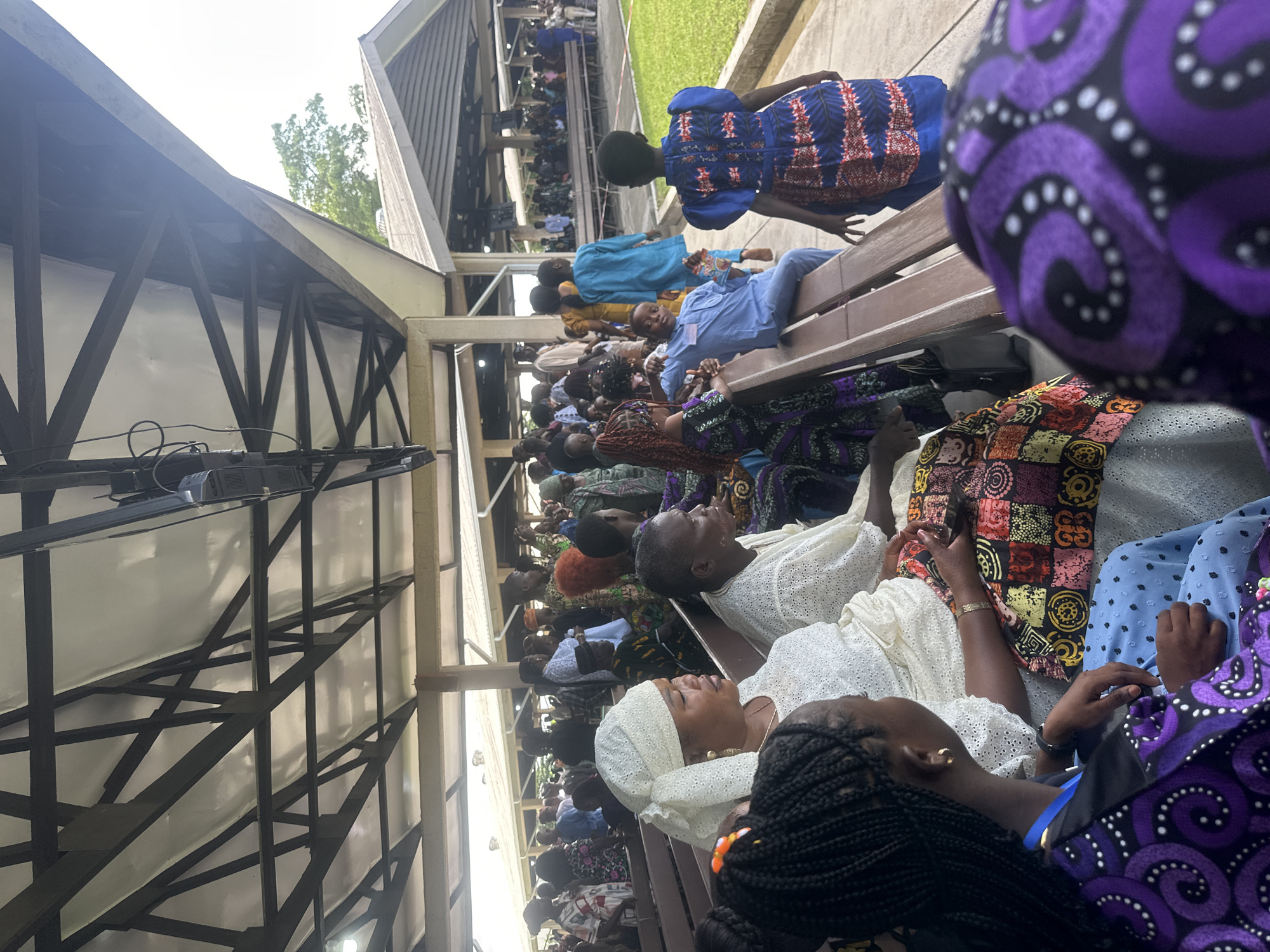 The image shows an outdoor gathering of people sitting on wooden benches under a covered roof supported by metal beams. The location is identified as Itoga, Nigeria. Many individuals are dressed in colorful traditional African attire. In the foreground, a woman wearing a white headscarf and white lace clothing is seated. To her left, a young girl with braided hair and a purple and black patterned outfit is also seated. To the right of the woman in white, a man with dark skin and white lace clothing is visible. Further in the background, more people are seated, and some are standing. The roof appears to be made of white material supported by dark metal structures. There is a grassy area visible to the right of the seating area. The lighting suggests it is daytime.