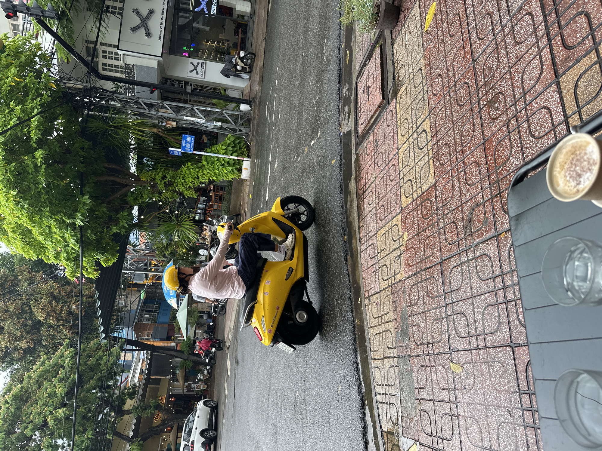 The image depicts a street scene, likely in Nha Trang Ward, Vietnam, showcasing a person riding a yellow scooter on a wet road. The rider is wearing a yellow helmet, a pink shirt, and dark pants. To the right of the scooter is a patterned sidewalk. In the background, there are trees, buildings, and street signs. The street signs include the name "Tô Hiến Thành." The building in the background has the logos "X" and "GOÛT SKR" and mentions the address "22 Tô Hiến Thành". A table with glasses and a cup is placed at the corner of the sidewalk. Cars and motorbikes are parked nearby.