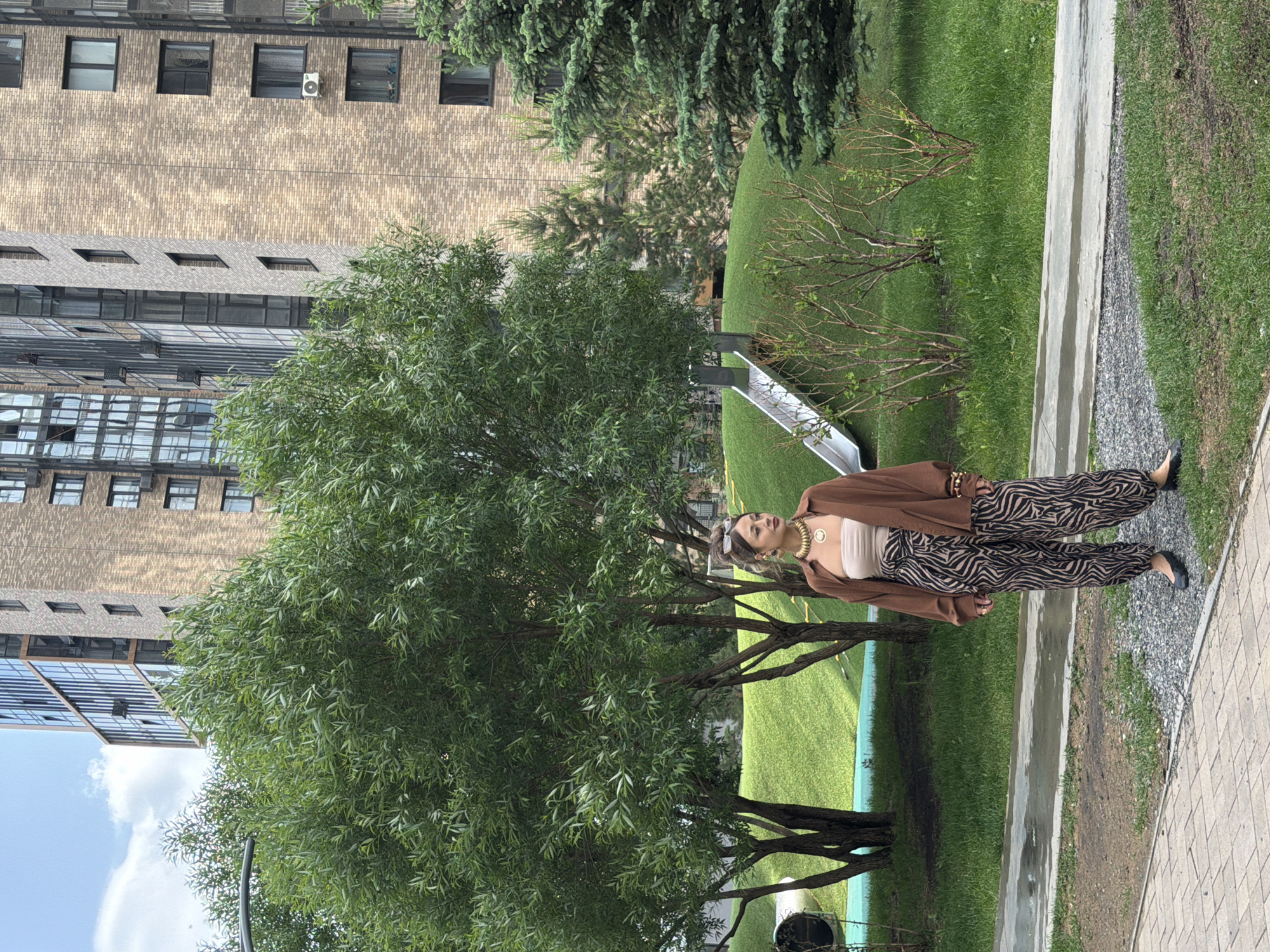 A woman stands on a paved walkway in an urban park, facing towards the right of the frame. She is wearing a light brown, open button-down shirt over a beige strapless top, and baggy zebra-print pants. Her accessories include a large gold necklace, hoop earrings, and sunglasses perched on her head. She is also wearing black ballet flats.
Behind her, a grassy hill slopes upward, with a silver slide descending from its peak. Several trees are scattered across the landscape, including a large, leafy tree to her left and a coniferous tree to her right. In the background, a modern brick apartment building with numerous windows rises several stories high. The scene appears to be located in the city of Krasnoyarsk, Russia, indicated by the architectural style of the building and the general aesthetic of the urban park. The weather is partly cloudy.
