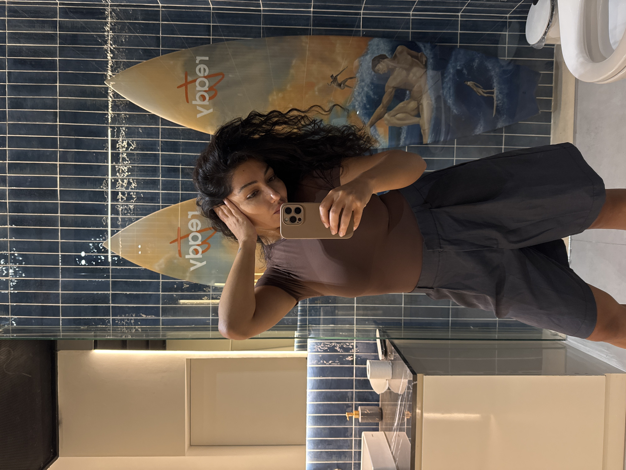 A woman is in a bathroom, taking a selfie with a smartphone. She has dark, curly hair and is wearing a brown shirt and gray shorts. The bathroom has dark blue tiled walls and two surfboards mounted on the wall with artwork. A toilet is visible in the upper right corner of the frame. The location is implied to be Kuta Selatan, Indonesia.