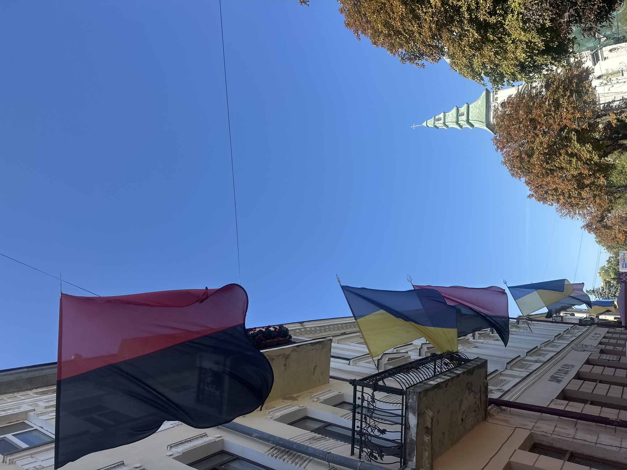 The image shows a street scene in Ternopil, Ukraine, with multiple flags hanging from a building. The flags are predominantly red, black, blue, and yellow. A balcony with an ornate railing is visible on the building, and several windows are present. Trees with autumn foliage are on the right side of the image. In the background, a church steeple with a green roof and a cross is visible. The sky is clear and blue. No people are visible in the image.