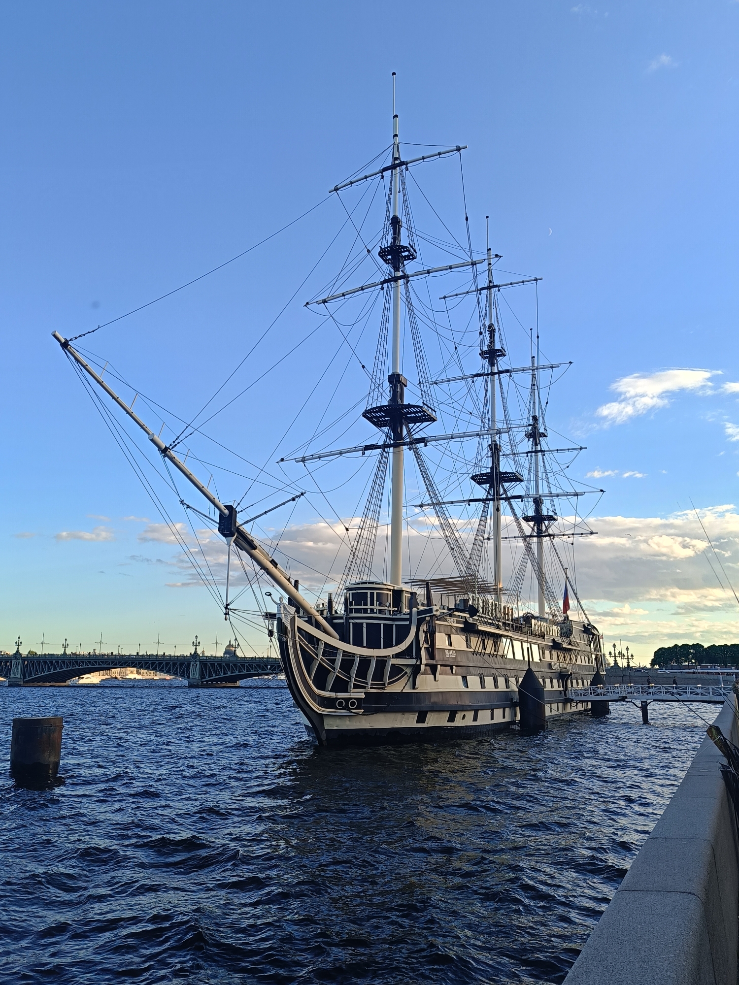 An anchored, three-masted sailing ship is docked on a body of water. The ship is dark with white trim and appears to be a historical replica. Its masts are tall and complex, with numerous ropes and rigging visible. A gangplank extends from the ship to the shore, where a stone embankment runs along the water's edge. In the background, a large bridge spans the water, and several buildings are visible on the distant shore. The sky is blue with some scattered clouds, and a crescent moon is visible. The water is dark and choppy. A single cylindrical post protrudes from the water near the ship.