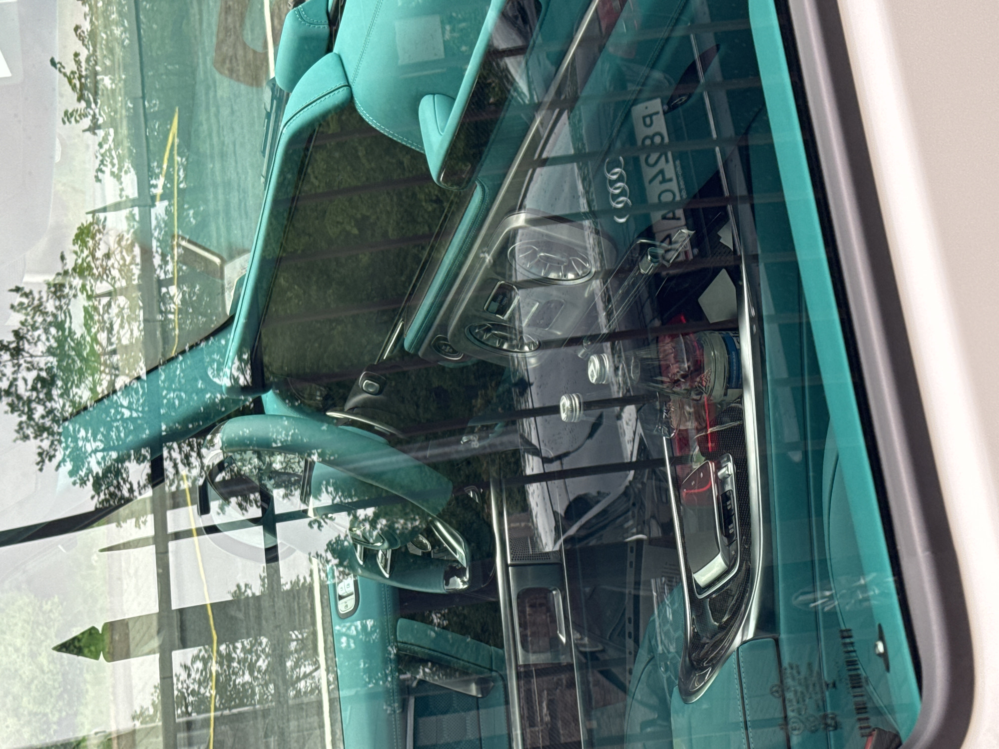 The image shows the interior of a car, likely an Audi, with turquoise upholstery, viewed through the rear window. Reflections in the window show trees and a building. A license plate is visible in reverse. Based on the reversed letters "APBSB9", and the presence of Audi rings, the image suggests the location is likely Saint Petersburg, Russia, as the license plate format matches those issued in that city. No people are visible in the image.