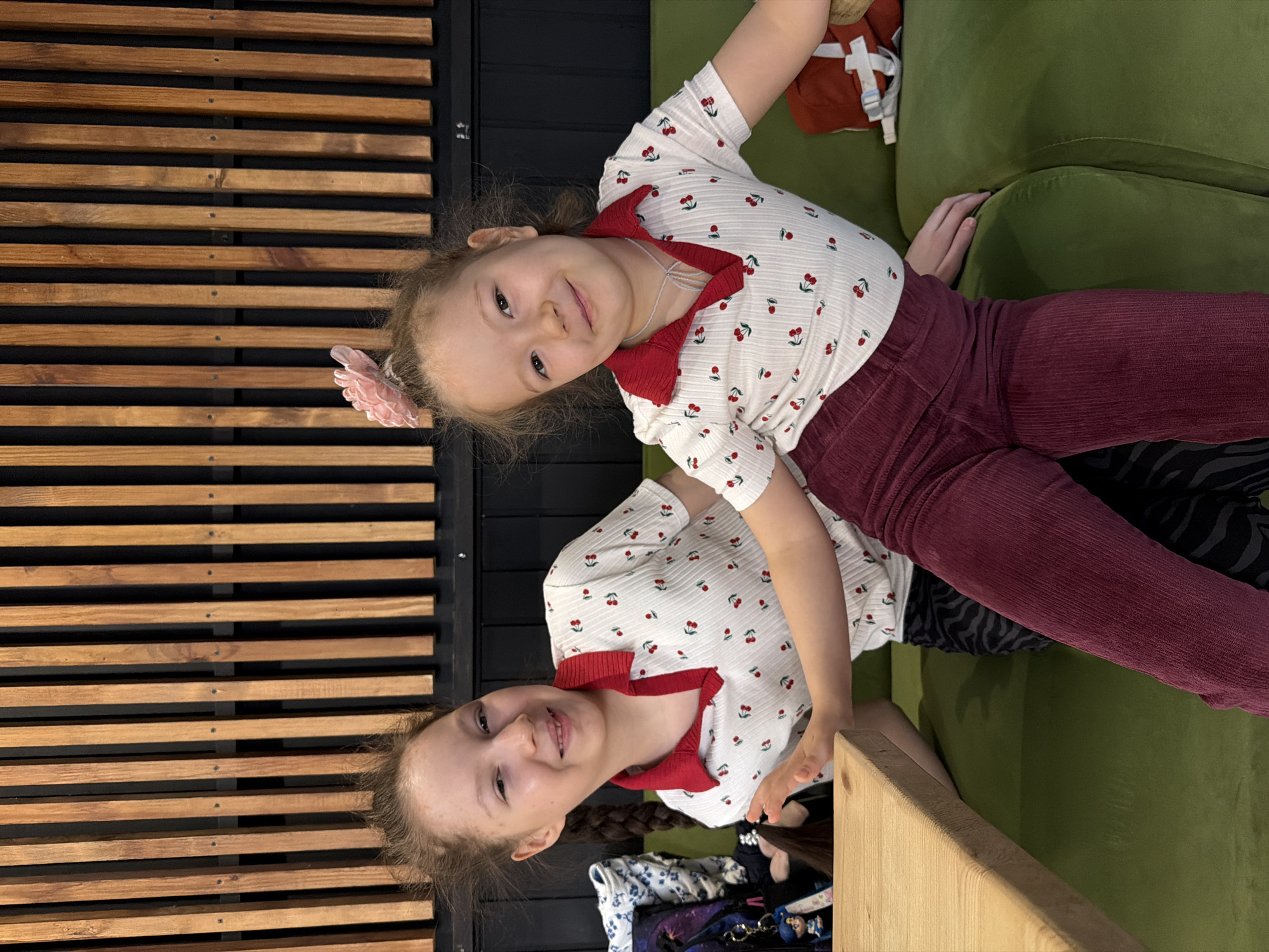 Two young girls are posed together on a green upholstered piece of furniture, likely a couch or sectional. The girls are wearing matching white shirts with red cherry prints and red collars, along with dark red corduroy pants. The girl on top has a pink flower clip in her hair. Behind the girls is a wall with horizontal wooden slats alternating with black strips. Personal belongings are visible in the lower portion of the frame, including bags and keys. The setting is indoors. The location of Izhevsk, Russia is not evident from the image.
