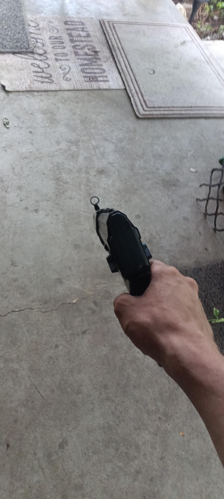 A person is holding a black pistol in their right hand. The pistol is pointed toward the upper-left corner of the image. The background includes a concrete surface, a doormat that says "Welcome to our homestead", and a second doormat. There are also glimpses of green vegetation and a metal grate. The location may be Coal Hill, United States of America.