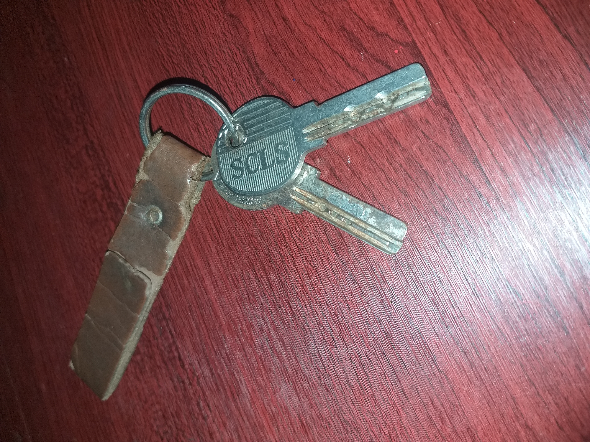 A pair of old, tarnished keys are attached to a worn leather fob by a silver keyring. The keys are made of metal and appear to be for a lock. The leather fob is brown and shows signs of age and wear, with creases and scuffs. The keyring is circular and silver-colored. The objects are lying on a red, wooden surface with a visible grain pattern. The background is a plain, dark red. There are no people or animals present in the image. The scene is static and there is no apparent action. The lighting is dim, creating shadows on the surface. The location is unknown and cannot be determined from the image.