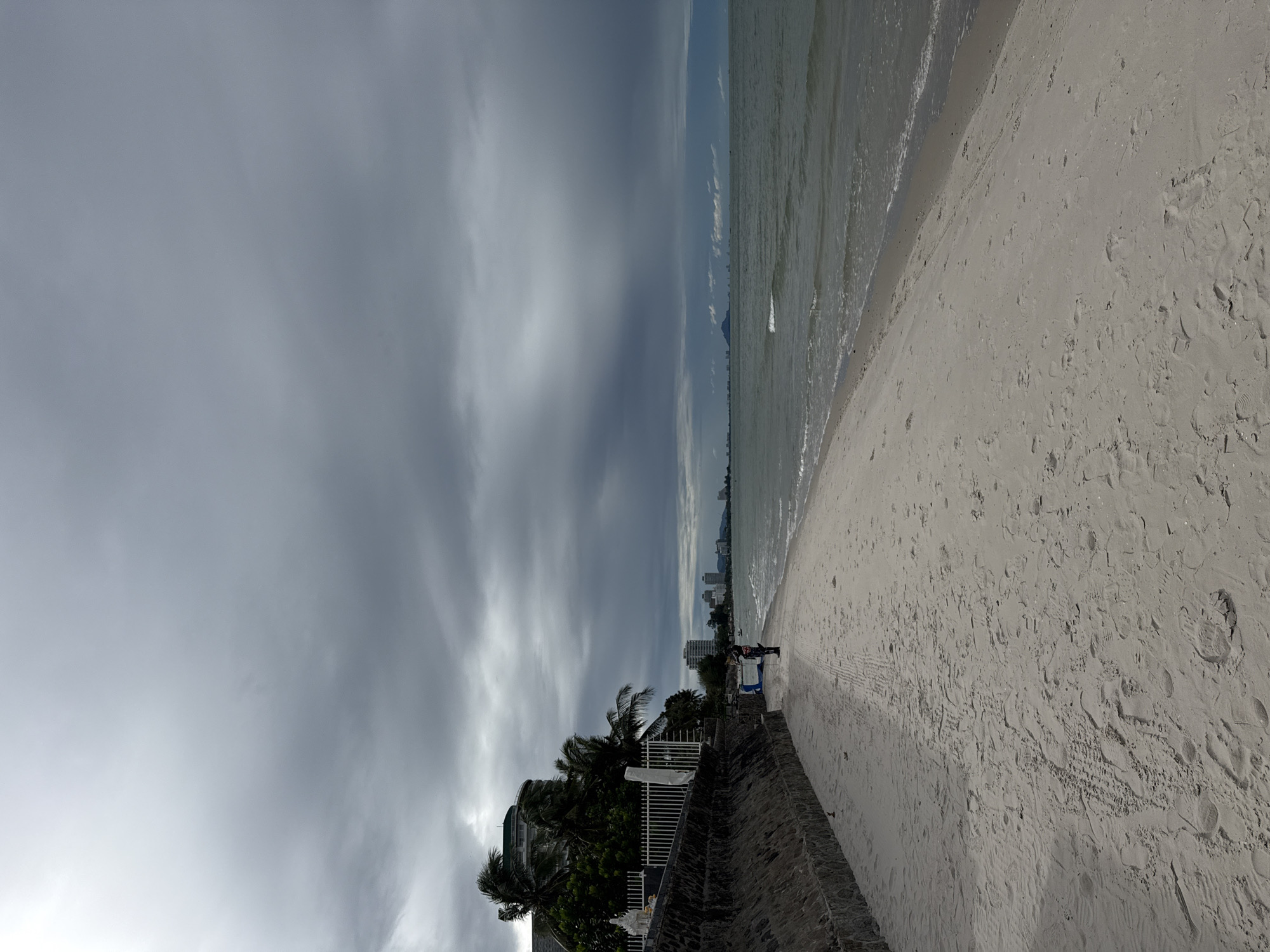 This is a vertical, eye-level, full shot of a beach scene in Hua Hin City Municipality, Thailand. The foreground is dominated by a wide, light-colored sand beach with numerous footprints and tire tracks. To the left, a textured stone wall runs along the edge of the beach, topped by a white fence. Behind the fence, several palm trees and a white, round building with a green roof are visible, suggesting a hotel or resort. In the mid-ground, two individuals, one wearing dark clothing and the other in lighter attire, are standing on the beach near the water's edge. The sea occupies the right half of the image, with moderate waves gently breaking on the shore. In the distance, a hazy skyline of buildings and a prominent, lone mountain rise from the horizon. The sky is heavily overcast with dark, dramatic clouds, creating a moody and atmospheric scene.