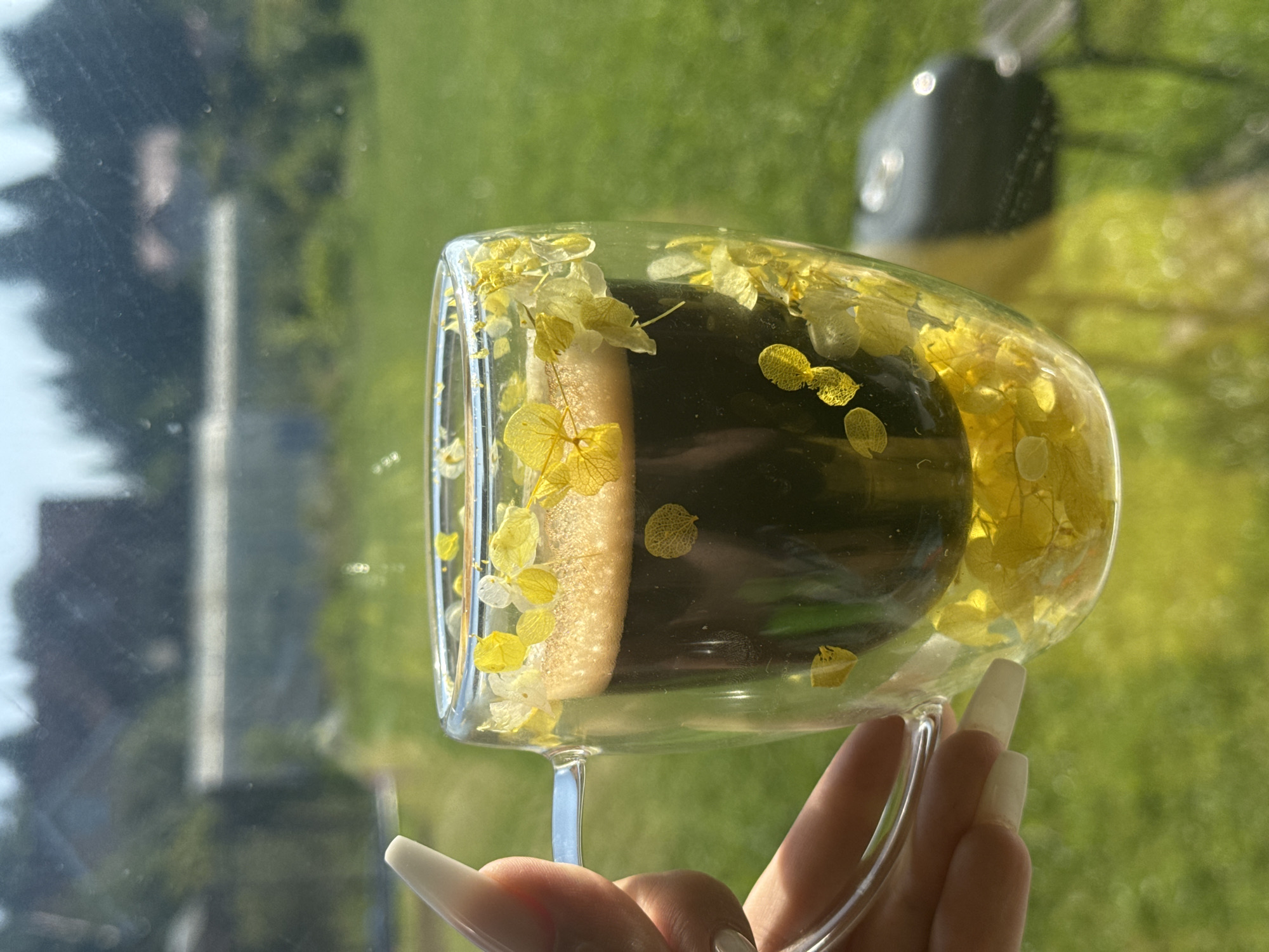 A close-up shot focuses on a clear, double-walled glass mug filled with dark coffee, topped with a creamy foam. The outer wall of the mug is adorned with small, dried yellow and white flower petals embedded within the glass. A hand with long, pale-colored artificial nails is holding the mug by its handle. The background is blurred, showing a green, grassy area with some out-of-focus foliage and structures. The lighting suggests it is daytime, with sunlight casting a warm glow on the scene. The location is identified as Shcheglovo, Russia.