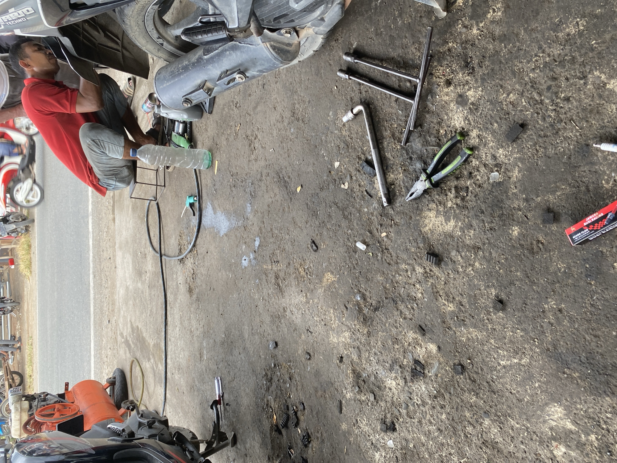 A man in a red shirt and grey pants is kneeling on a small stool in front of a motorcycle. He is working on the motorcycle. Tools are scattered on the ground around him, including a wrench, pliers, and a bottle of water. An orange air compressor is visible in the background, as well as a street and other motorcycles. The ground is dirty and appears to be a workshop or garage.