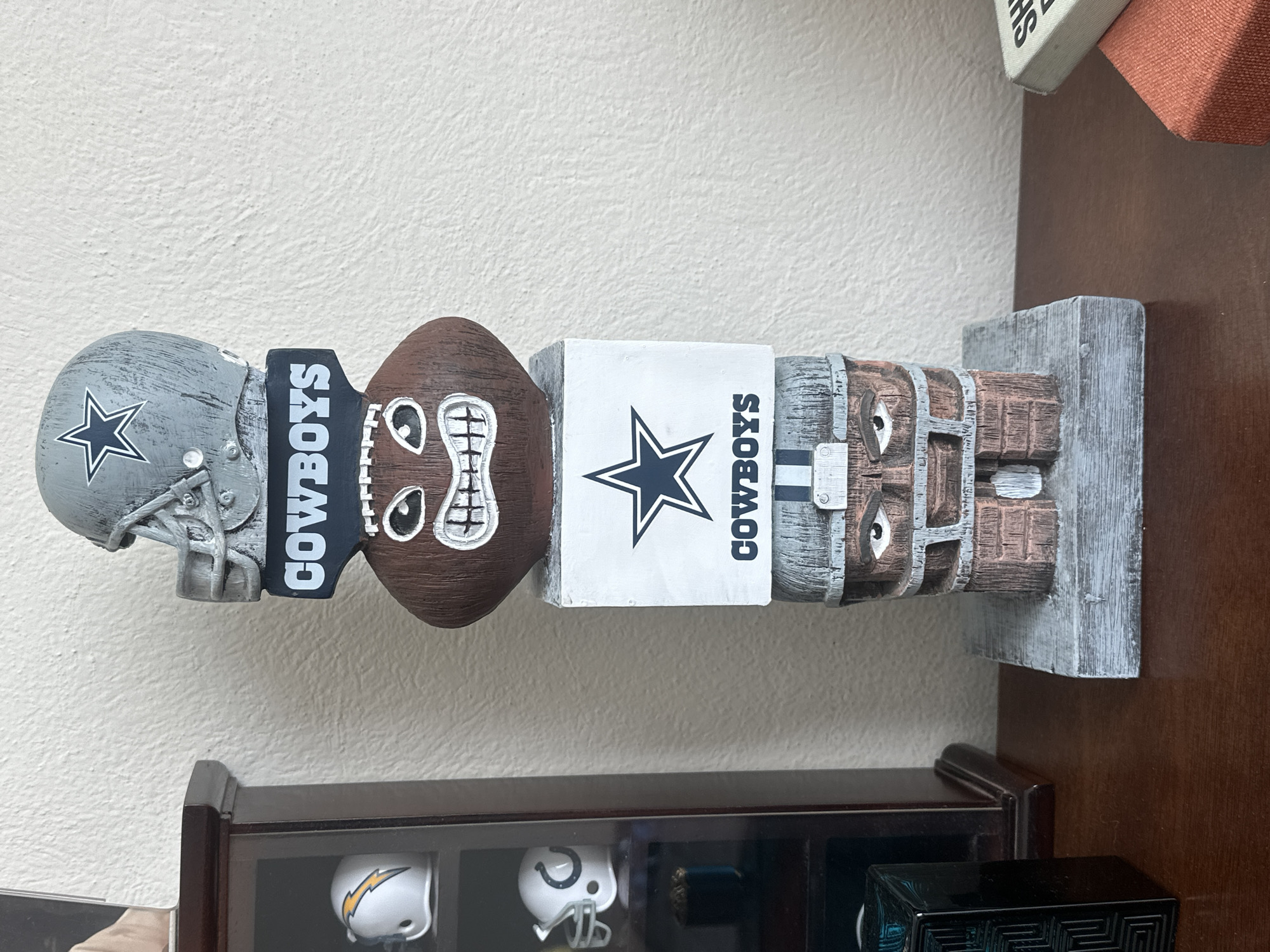 The image shows a Dallas Cowboys totem pole resting on a dark wood shelf. The totem pole consists of a Dallas Cowboys helmet, a "Cowboys" nameplate, a football with a face, another "Cowboys" nameplate with the team star logo, and a stacked log base with two eyes. The shelf also contains display cases with mini NFL helmets. The location is Jesús del Monte, Mexico.