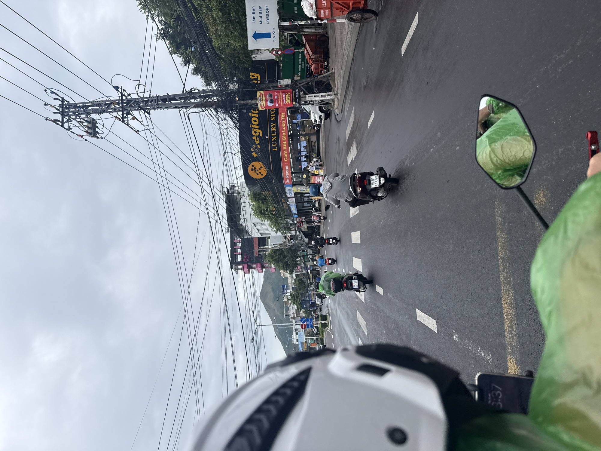 A first-person perspective, looking forward from a motorcycle in Nha Trang, Vietnam, shows a wet asphalt road under a cloudy sky. Multiple motorcycles and scooters are on the road, some with riders wearing raincoats. Buildings line the street, including a large store with a prominent yellow and black sign that reads "thegioididong LUXURY STO." A traffic light is visible, as are power lines crisscrossing overhead. In the distance, a mountain is visible. The phone mounted to the motorcycle shows the time as 3:37. The street is wet, indicating recent or ongoing rain.