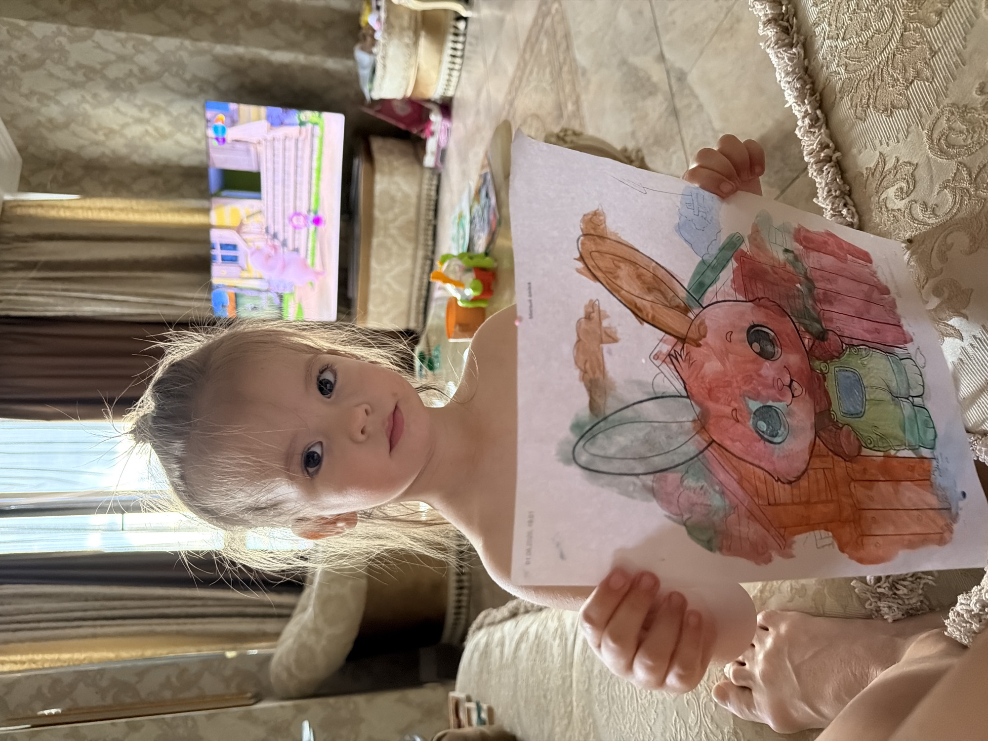 A young girl with blonde hair is holding up a colorful drawing of a rabbit. She is looking at the camera with a curious expression. The drawing appears to be watercolor and crayon. The girl is sitting on a couch with a textured fabric. In the background, there is a television screen displaying a cartoon, and various objects on shelves and tables. The scene suggests a home environment, likely a living room. The lighting indicates it is daytime. The image has a casual and candid feel, capturing a moment of the girl showing off her artwork. The location is Ufa, Russia. There is no visible text in the image, except for what appears to be a timestamp on the drawing itself.