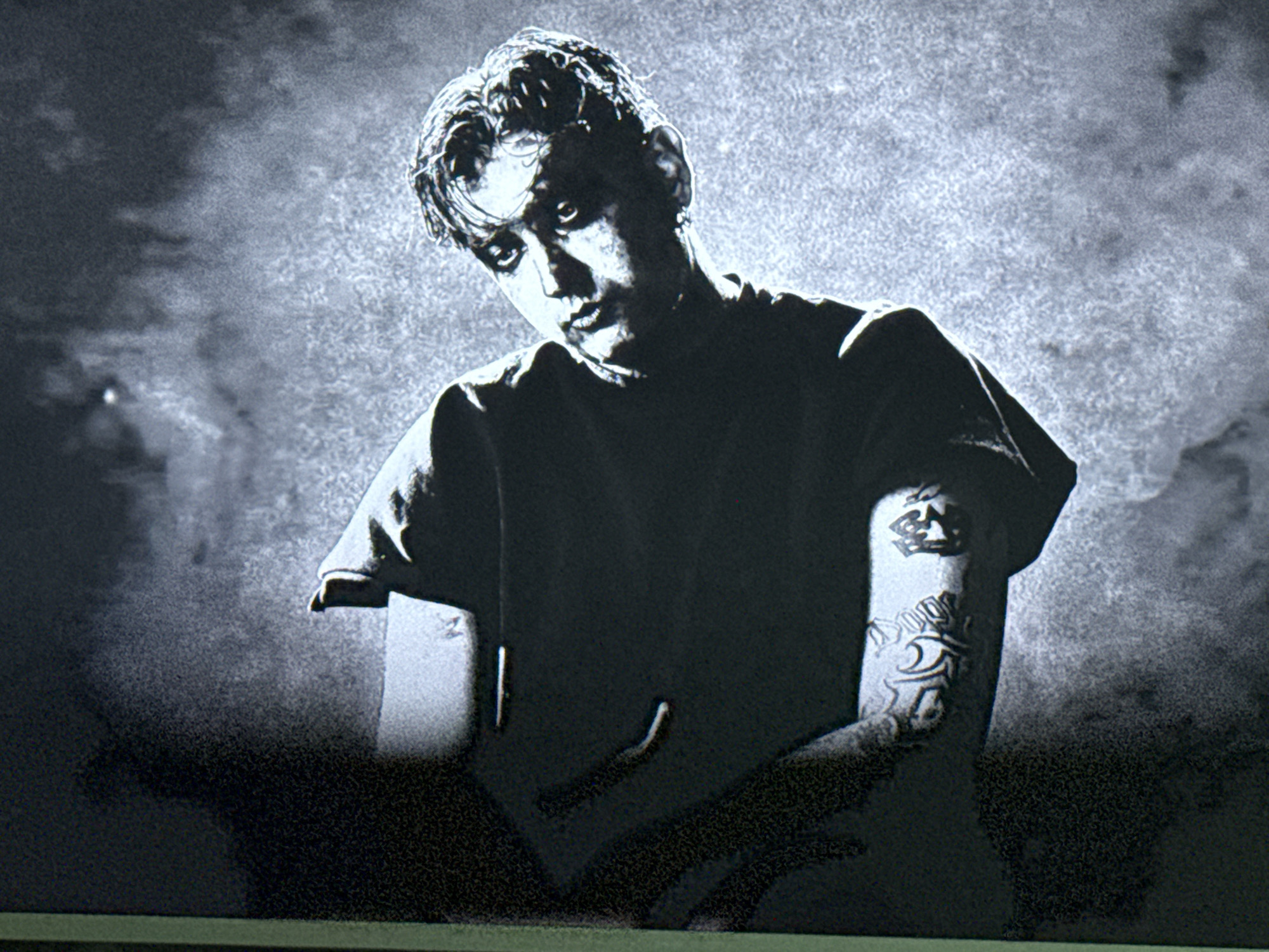 A medium shot shows a man in a dark t-shirt with his arms bent in front of him. The man has fair skin, with dark hair and dark eyes, he is looking slightly down and to the left. The man has tattoos on his left arm, including the image of a crown and lettering. The background is a grainy off-white. The location is Unknown, Unknown.