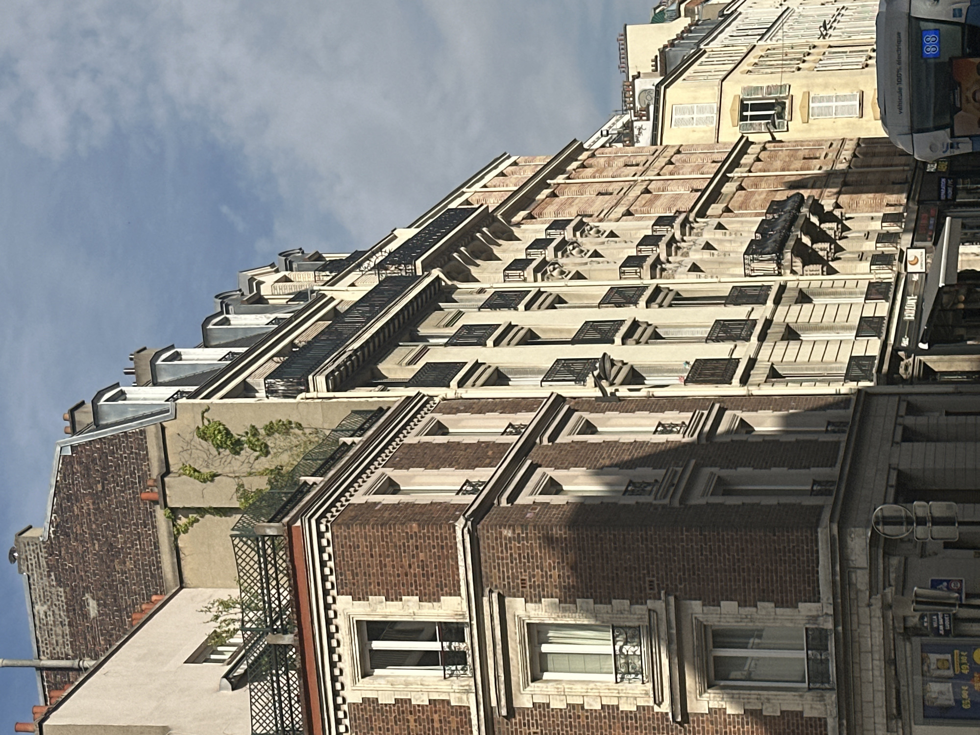 The image depicts a street view of Parisian architecture on a sunny day with scattered clouds. The foreground features the facade of a multi-story building constructed with dark red brick and ornate stone trim around the windows and corners. The building has several floors with rectangular windows, some of which are French doors leading to small balconies with black iron railings. Above these, a higher floor showcases dormer windows and a rooftop terrace with a decorative railing and some vegetation.
Adjacent to this building, to the right, are other buildings with similar architectural styles, characterized by balconies, large windows, and pitched roofs with dormers. The buildings are tall and closely packed, typical of Haussmannian architecture found in Paris. The facades vary in color, with some being light cream or beige, and others, like the one in the immediate foreground, being brick.
In the lower right corner of the image, the back of a modern electric bus is visible. The bus is light grey with "véhicule 100% électrique" written on its side, along with a blue number "88" displayed prominently. Some street signs and storefronts are also partially visible at the bottom of the frame. The overall impression is a classic Parisian street scene, likely in an urban residential or commercial district.
