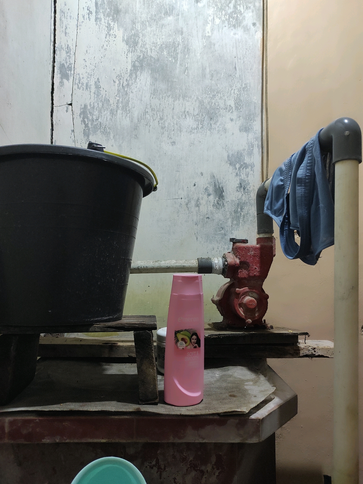 The image depicts an outdoor or semi-outdoor scene, possibly a utility area. In the foreground, a large black bucket sits on a rustic wooden platform. To the right of the bucket, a small red water pump is visible. A blue garment, possibly underwear, is draped over a PVC pipe connected to the pump. A pink bottle, possibly shampoo or conditioner, stands in front of the pump. The background features a cracked and weathered concrete wall. The overall environment appears utilitarian and somewhat unkempt, with a focus on functional objects. The scene is lit by artificial light, casting shadows. The location is identified as Unknown, Unknown.