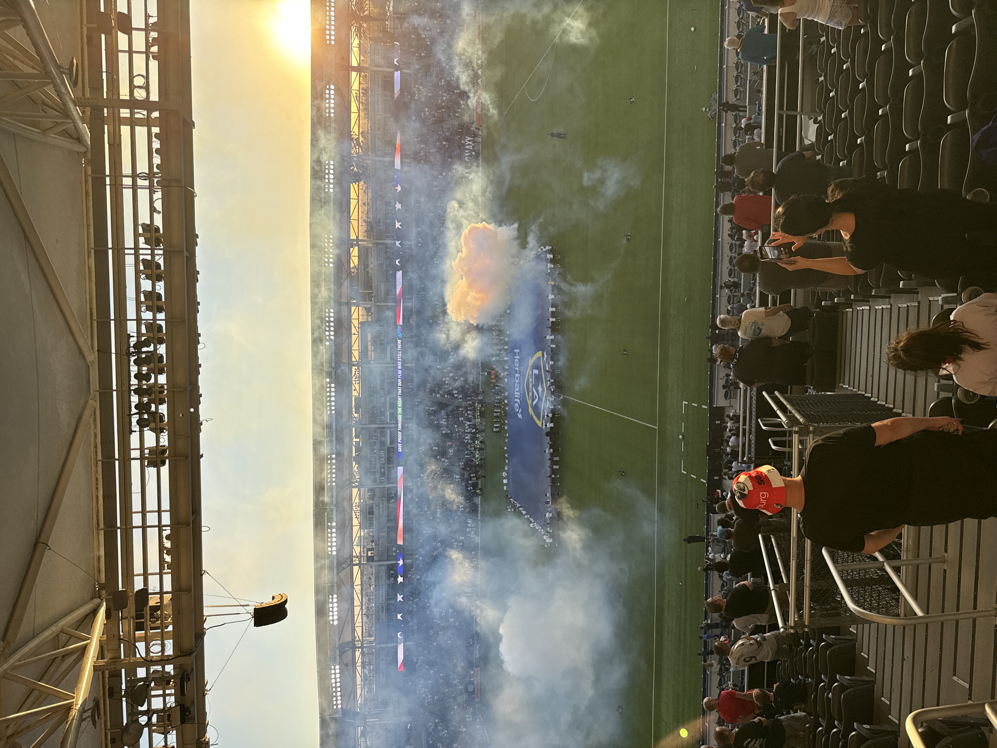 The image depicts a wide view of Dignity Health Sports Park in Carson, California, during what appears to be a pre-game or halftime ceremony. The sun is setting, casting a warm glow across the stadium. A large banner is unfurled on the field by a line of people, and colored smoke is being released, creating a dramatic visual effect. The stands are partially filled with spectators, some of whom are holding up their phones to capture the moment. The stadium features tiered seating, stadium lights, and a green soccer field.
