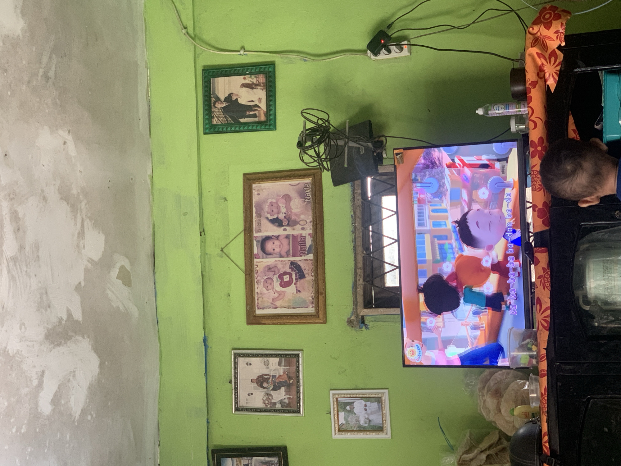 The image depicts an indoor scene, likely a living room, with vibrant green walls. The ceiling shows signs of wear and discoloration. Several framed photographs are hung on the wall, including what appear to be family portraits and baby pictures. In the foreground, a television screen displays a cartoon. Various items are placed on a shelf beneath the television, including what seems to be a small collection of toiletries and other household objects. A young child is visible from behind, facing the television. The overall impression is that of a simple, lived-in domestic space. While the image does not contain explicit clues to its exact location, the general aesthetic and objects suggest a residence in a developing country. Based on the provided information, the image could be located in Palembang, Indonesia.