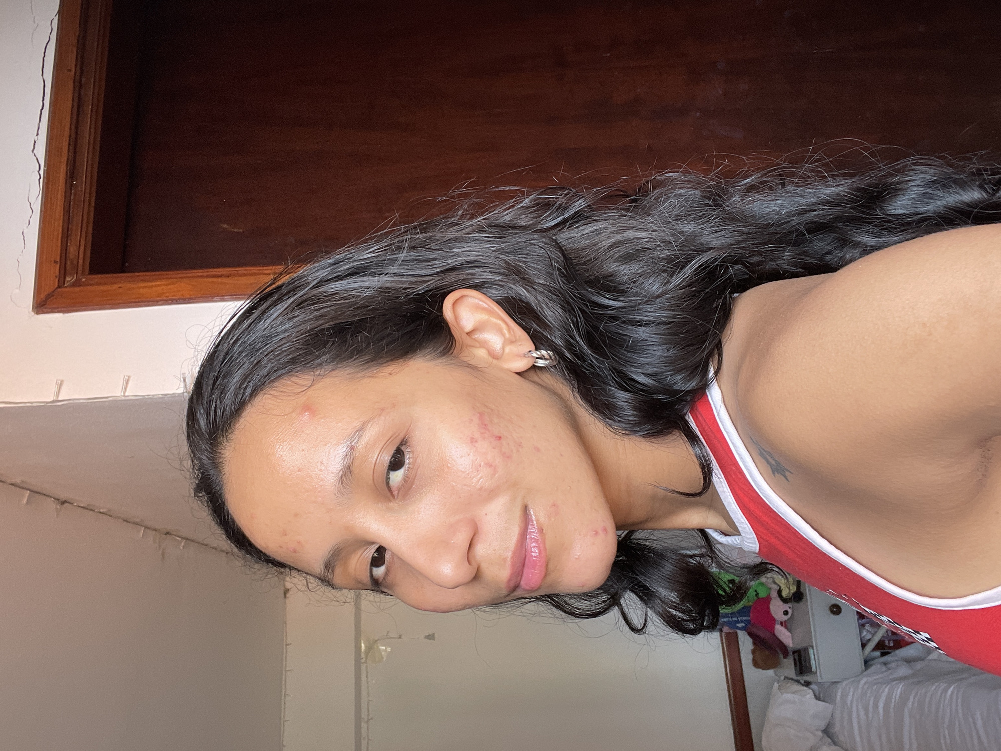 The image shows a young woman with dark, wavy hair, wearing a red and white tank top. She has several pimples on her face, particularly on her cheeks and forehead. She is looking towards the camera with a slight smile. The background appears to be an indoor setting, with a wooden door and a wall with some visible cracks. There is a bed with white linens and some stuffed animals visible on the left side of the image. The lighting suggests it is daytime. The location is specified as Porlamar, Venezuela.