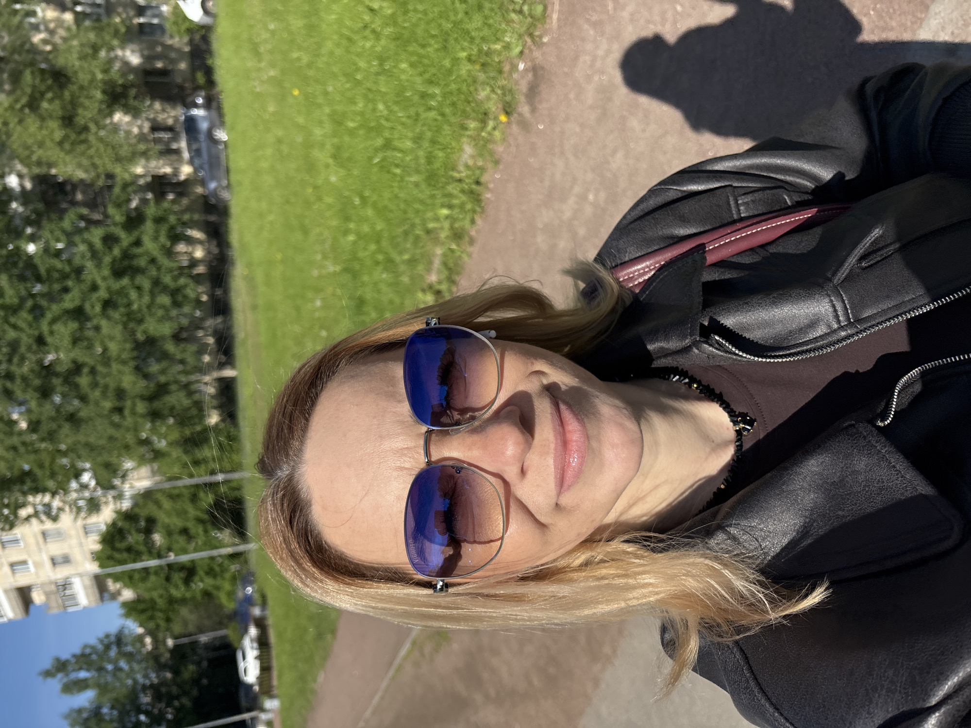 The original image, rotated ninety degrees clockwise, captures a selfie of a woman outdoors on a bright, sunny day. The subject is a woman, appearing middle-aged, with blonde hair that frames her face. She is wearing a black leather jacket and large, reflective blue sunglasses with silver frames, which obscure her eyes. Her lips are slightly parted, forming a gentle, subtle smile. A dark, possibly beaded, necklace is visible around her neck.
The background shows an outdoor setting with a paved path or road beside a vibrant green grassy area. Several trees are scattered in the mid-ground, and in the distance, parts of a multi-story building can be seen. Multiple cars are parked along what appears to be a street. The bright sunlight casts distinct shadows on the ground, indicating clear weather.
Visible text on the bottom left lens of the sunglasses reads "VIRTUAL-O". The scene suggests an urban or suburban environment, possibly a park or a street. The location is unknown, Russia.