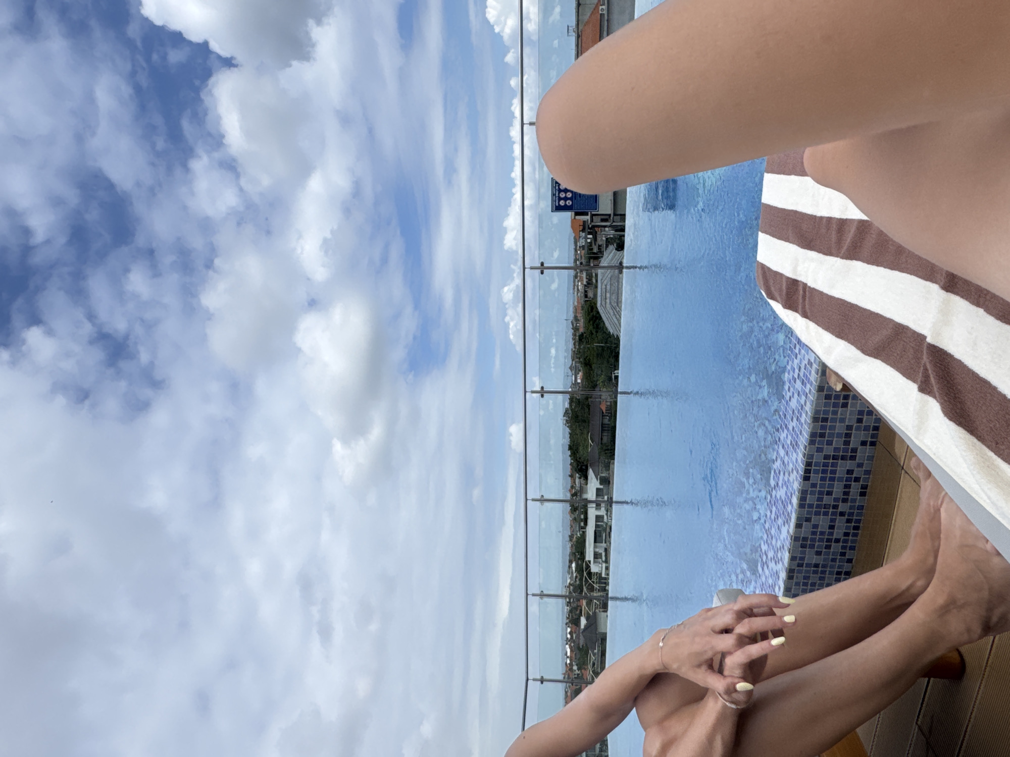 The image depicts a rooftop infinity pool with a view of North Kuta, Indonesia. The sky is filled with scattered clouds. In the foreground, a person's legs and hands are visible, resting on a striped lounge chair. The person's hands are clasped, and they are wearing a bracelet. The pool water is a vibrant blue, and the edge of the pool is tiled with blue and white mosaic. Beyond the pool, a cityscape with houses and trees can be seen, extending towards the horizon. A glass railing separates the pool area from the cityscape.