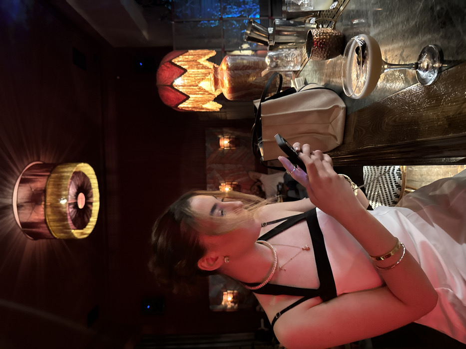 The image displays an indoor bar or lounge in Yerevan, Armenia. The specific name or address of the establishment is not visible. An adult female is seated at a dark, veined bar counter in the left foreground, her gaze directed downwards at a black smartphone she holds with both hands. Her dark brown hair is styled up, with some strands loose around her face. She wears a white top with black halter and spaghetti straps. Jewelry includes a gold-colored choker, two gold-colored pendant necklaces (one with a 'T' shaped charm, the other a small animal or charm), small stud earrings, two rings on her left hand, and multiple gold-colored bangles on her right wrist.
On the counter before her, a coupe glass holds an amber-colored cocktail garnished with two dark objects on a pick. To her right, a light beige tote bag with black straps, a hammered copper mug, and a clear glass with ice are visible. Behind these, stainless steel bar tools are present.
The environment is characterized by dim, warm, reddish-orange lighting. A two-tiered cylindrical ceiling lamp with red and gold fabric is suspended overhead. A large table lamp with a red, fringed shade and an ornate base sits on the counter in the middle ground. The background wall is dark red, featuring two illuminated wall sconces. A blurred male figure is partially visible in the mid-background, wearing a light-colored shirt. A textured, possibly tiled or glass block, wall section on the far right is illuminated with a blue light.