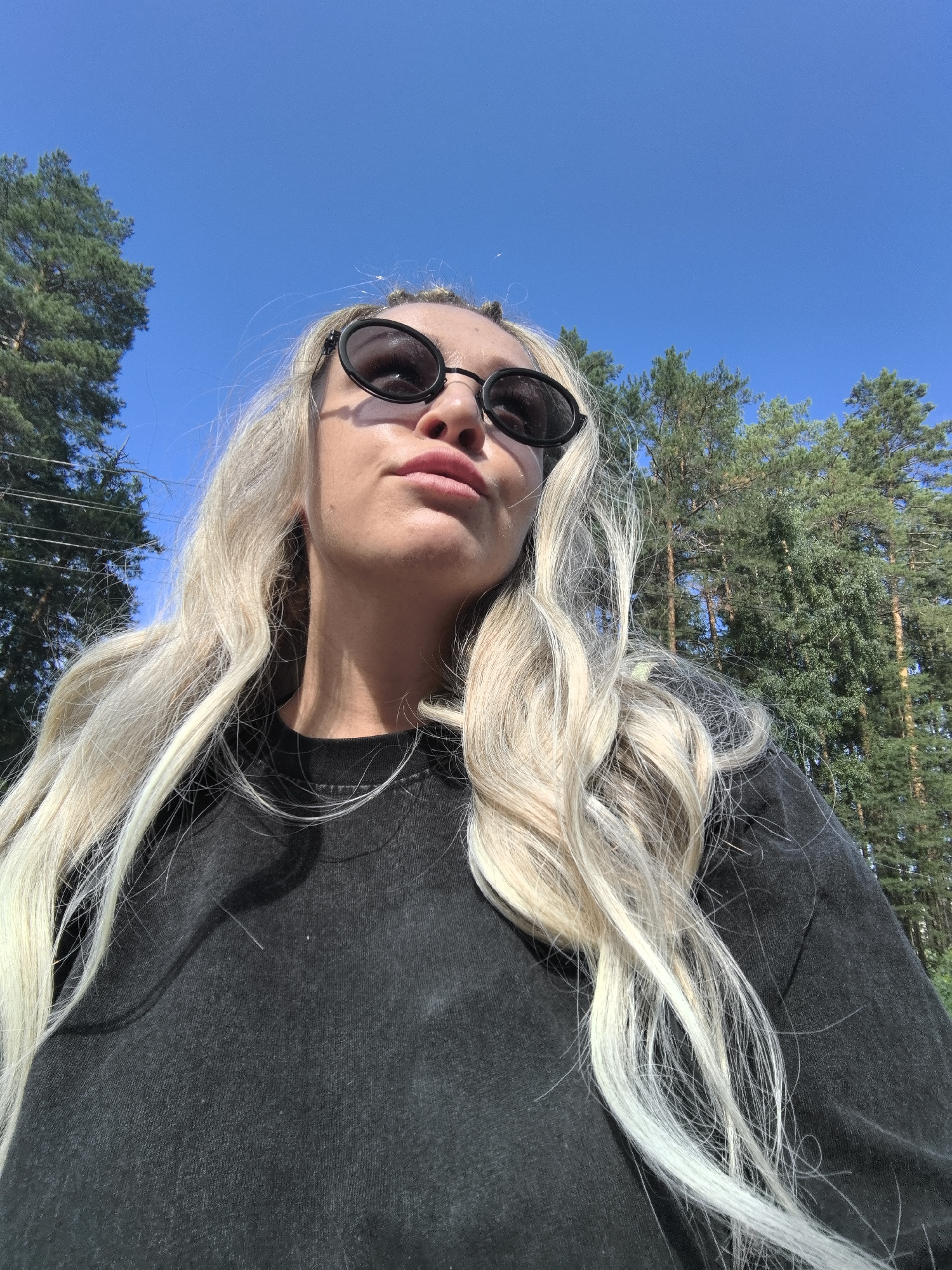 A selfie, taken from a low angle, shows a woman with blonde hair and sunglasses against a clear blue sky and a backdrop of pine trees. She is wearing a dark t-shirt. The lighting suggests it is daytime. The location is specified as Noviy, Russia.