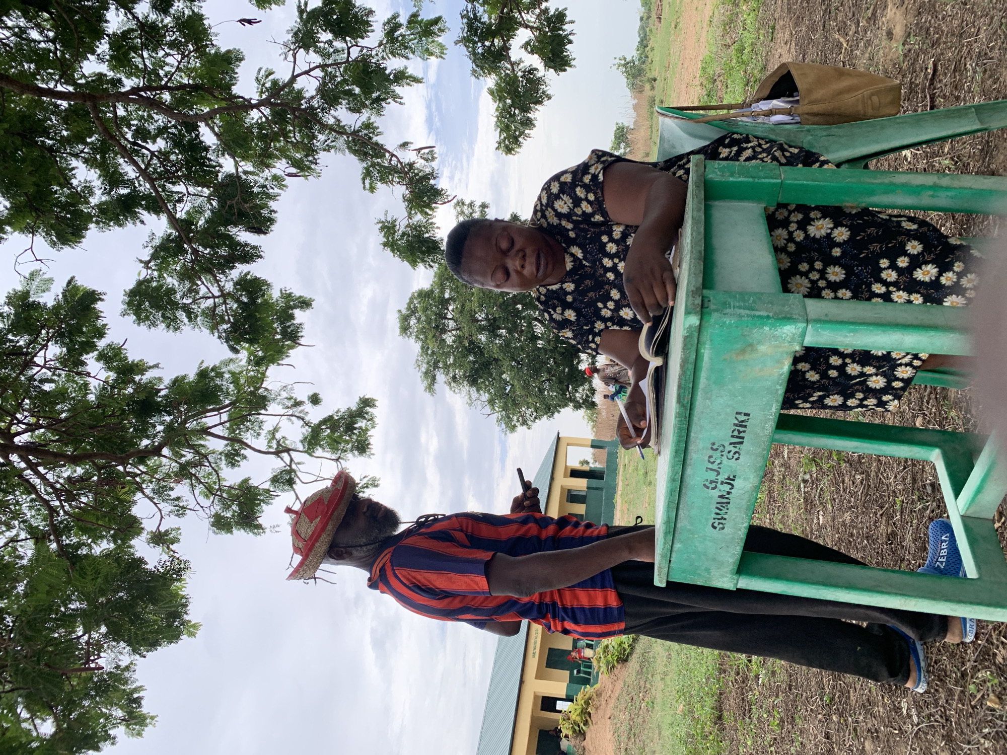 This image captures an outdoor scene in Akwanga, Nigeria, showing two individuals engaged in an activity, likely educational or administrative, under the shade of a large tree.
In the foreground, a woman is seated at a green plastic desk, wearing a black dress adorned with white daisy patterns. She is looking down, seemingly engrossed in reading or writing in a book or notepad. A brown shoulder bag rests on the back of her chair. Opposite her, a man stands, wearing a striped red and blue short-sleeved shirt, dark pants, and a distinctive straw-like hat with red trim. He has a beard and is holding a smartphone in one hand and appears to be writing or making notes in a small pad with the other. He wears bright blue shoes with the word "ZEBRA" visible on them.
The setting is outdoors, with the canopy of a large tree overhead, providing shade from what appears to be a bright, overcast day. In the background, a light yellow building with a dark green roof, resembling a school or public facility, is visible. The ground is a mix of dry grass and dirt.
Visible text on the side of the green desk clearly reads: "G.J.S.S. GWANJE SARKI". This likely refers to a school, possibly "Government Junior Secondary School Gwanje Sarki." The man's shoes also display the brand name "ZEBRA". The overall atmosphere suggests a focused, informal learning or work environment taking place outside.