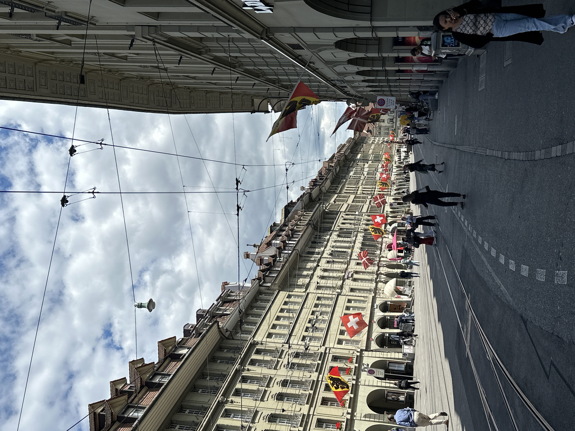 The image depicts a wide street in Bern, Switzerland, lined with traditional European architecture. The buildings on either side are multi-story with arched ground floors and numerous windows. Swiss flags are prominently displayed, hanging from poles along the facades of the buildings on the left side and from wires spanning the street. The street itself has tram tracks and is paved with asphalt. Several individuals are walking on the sidewalks and crossing the street, dressed in casual attire. The sky is partly cloudy, casting shadows on the street. On the right side of the street, the buildings are also of similar architecture, with shops visible at ground level, some with awnings. Overhead, electrical wires for what appears to be a tram system are strung across the street.