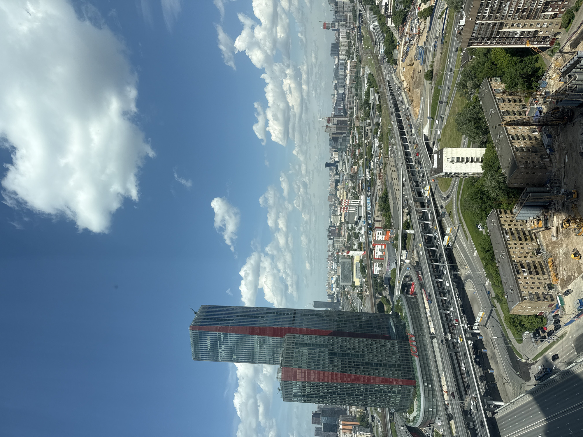 This image captures a high-angle view of a sprawling urban landscape in Khimki, Russia, bathed in bright midday sunlight under a clear blue sky with scattered white cumulus clouds.
In the foreground, a prominent modern skyscraper with dark glass facades and distinctive red horizontal stripes stands tall. Below and around it, a complex and extensive network of multi-lane highways, elevated roads, and railway tracks crisscrosses the scene. Numerous vehicles are visible on the roads, indicating active traffic and urban movement.
The cityscape extends into the midground and background, featuring a dense array of residential and commercial buildings of varying heights. Several areas show signs of ongoing development, with construction sites marked by cranes, exposed building materials, and earthmoving equipment. Green trees and patches of grass are interspersed throughout the concrete environment, particularly along roadsides and between buildings.
Notable visible text includes "МГИМО" (MGIMO) and "МЕГА" (MEGA), displayed in red letters on buildings situated near the main highway interchange. The overall scene portrays a dynamic, modern city characterized by its extensive infrastructure and continuous growth. No specific events or individual emotions are discernible.