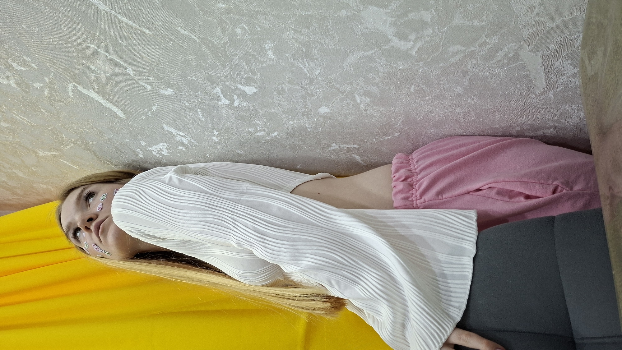 A young woman with long blonde hair is shown lying horizontally on her back, looking upwards. Her hair spreads out on a vibrant yellow surface, possibly a blanket or cushion. Small, decorative stickers, resembling tiny flowers, are applied to her face near her eyes. She wears a white, long-sleeved top with a distinct pleated or ribbed texture, revealing a narrow strip of her midriff. Her lower body is dressed in pink, loose-fitting sweatpants with an elasticized waistband.
The background consists of a wall covered in light-colored, possibly off-white or very light grey, textured wallpaper that features an abstract, mottled pattern. On the right side of the frame, a dark grey piece of furniture, likely a sofa or chair, is partially visible. The lighting is soft and ambient, suggesting an indoor setting. The woman's expression is neutral and calm, indicating a relaxed state. No specific activity other than posing or resting is apparent. There is no visible text in the image. The scene is set indoors in Chaikovsky, Russia.