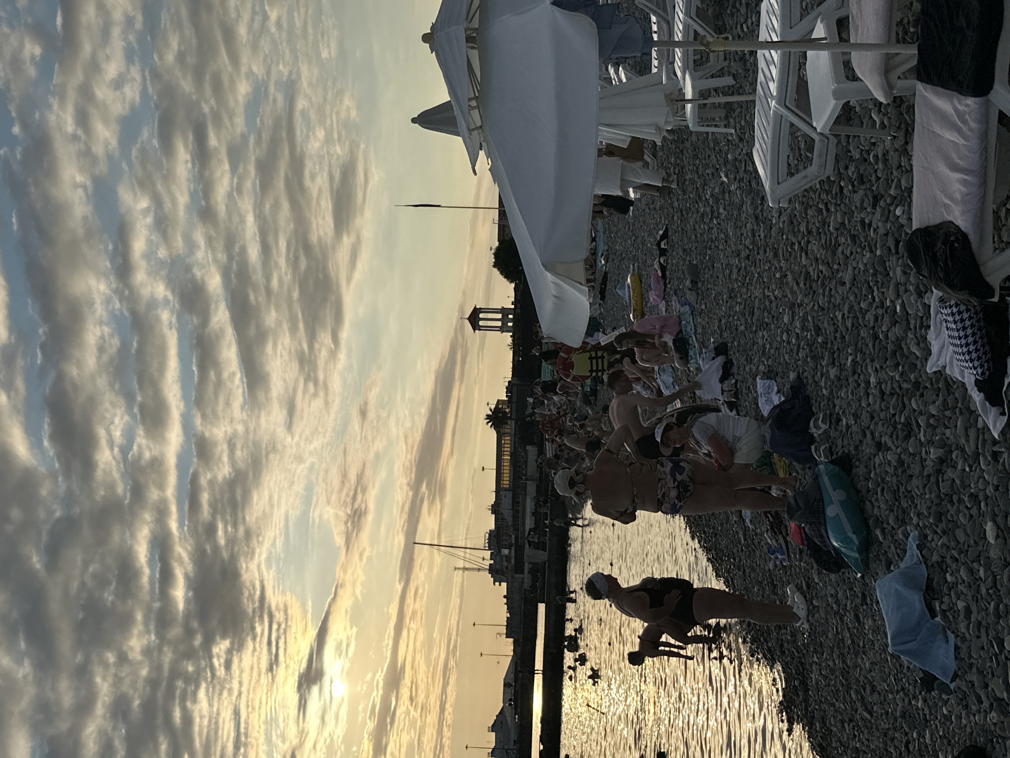 The image depicts a pebble beach on a cloudy day, with the sun partially visible through the clouds, casting a warm glow on the scene. The sea is calm, and several people are wading in the water or standing on the shoreline. On the beach, a crowd of people are seen relaxing, with some sitting on towels and others lying on lounge chairs. Beach umbrellas are set up, providing shade. In the background, a harbor with boats and buildings is visible, suggesting the location is a coastal city. The overall atmosphere is that of a leisurely summer day at the beach. Given the architecture of the tower in the background and the general ambiance, this scene is likely located in Sochi, Russia, possibly near the port area or one of its popular beaches.