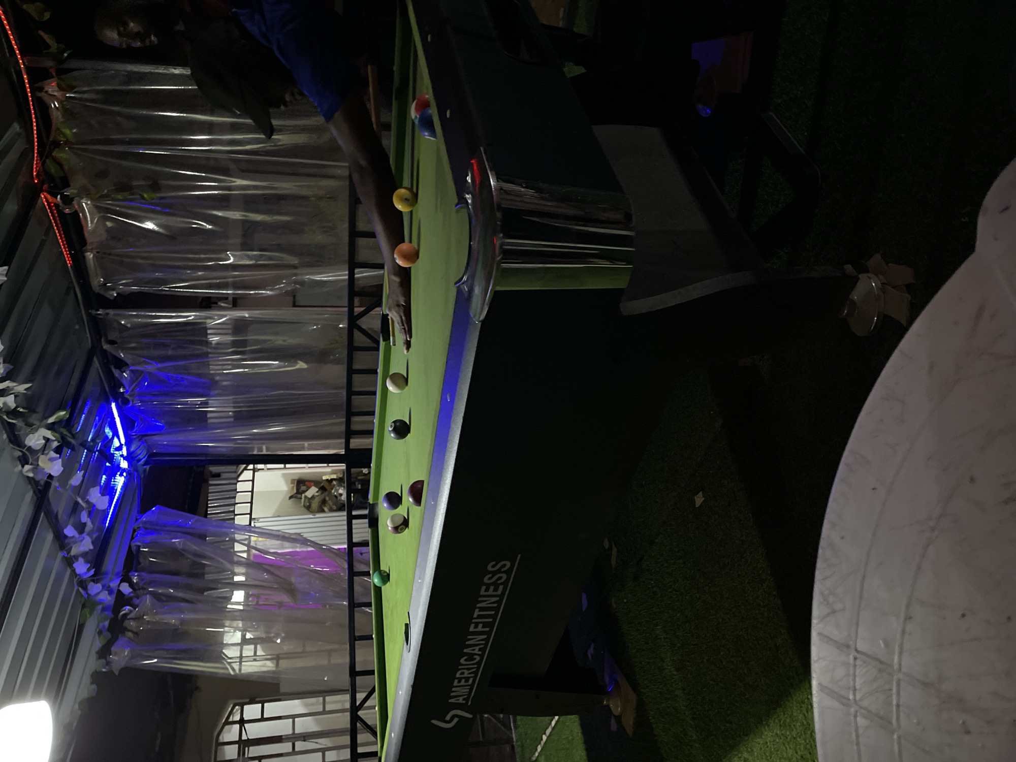 A person is playing pool on an "American Fitness" brand pool table. The scene is set outdoors, possibly in a backyard or patio area, in Barakin Akawo, Nigeria, based on image information. The lighting is dim, with plastic sheeting adorned with white flowers and colored lights partially enclosing the space. A white table is present in the lower right of the image, while the pool table is covered with pool balls.
