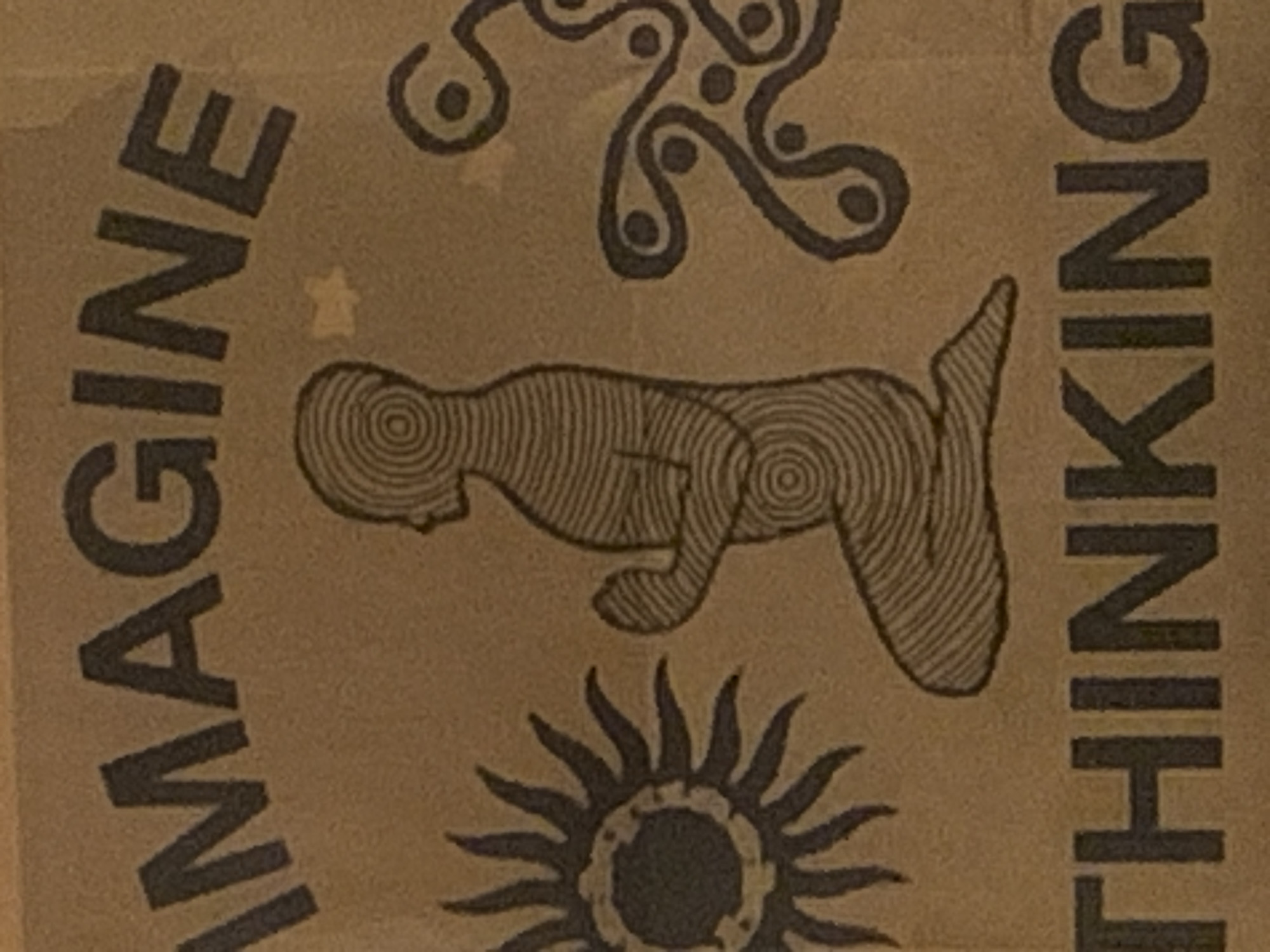 The image shows a close-up of a printed design on a brown surface, possibly paper or fabric. The design features stylized illustrations and text. There is a stylized human figure drawn with concentric circles filling its form. Below the figure is an image resembling a sun with radiating rays. Above is an abstract blob-like shape made up of interconnected circles. The text "IMAGINE" is vertically aligned on the left and the word "THINKING" is vertically aligned on the right, sandwiching the illustrations. The overall style has a hand-drawn, slightly rough quality. There is no explicit indication of Barcelona or any specific location in Spain.